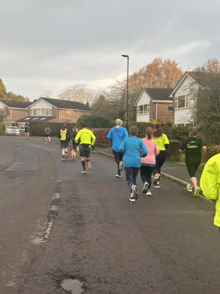 Group of people jogging or walking on a neighborhood street, some wearing high-visibility clothing, with houses and autumn trees in the background.