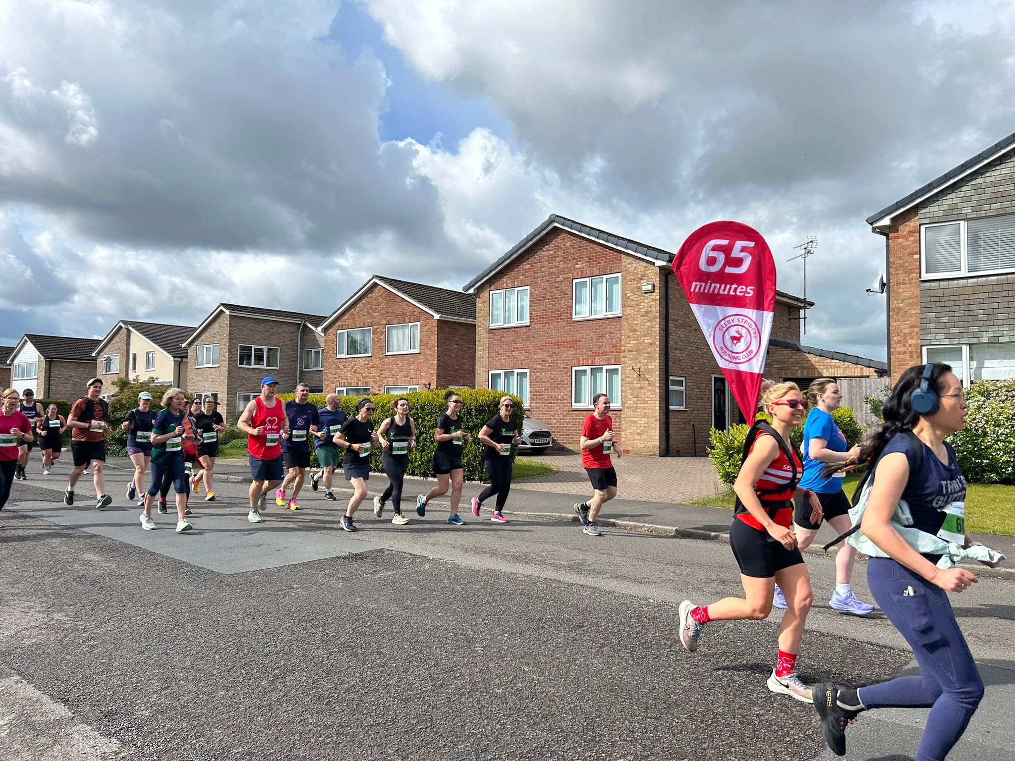 A large group of people participating in a running event outside residential houses, with a red flag that reads '65 minutes' and a logo for a running club. The sky is partly cloudy.