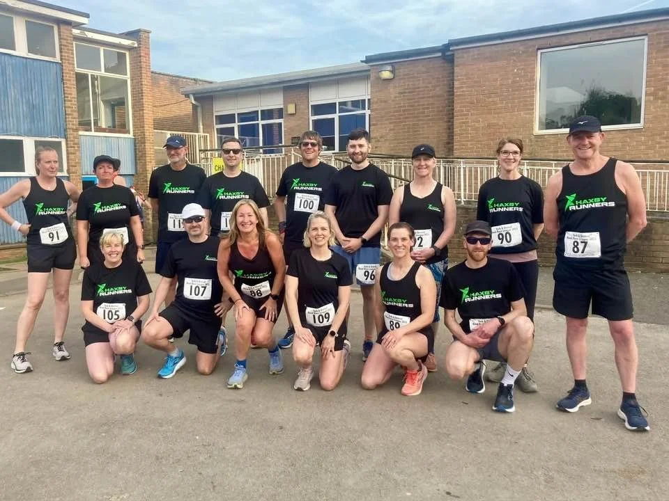 Group of runners in black athletic wear standing and kneeling outdoors with residential buildings in the background, posing for a photo before a race.