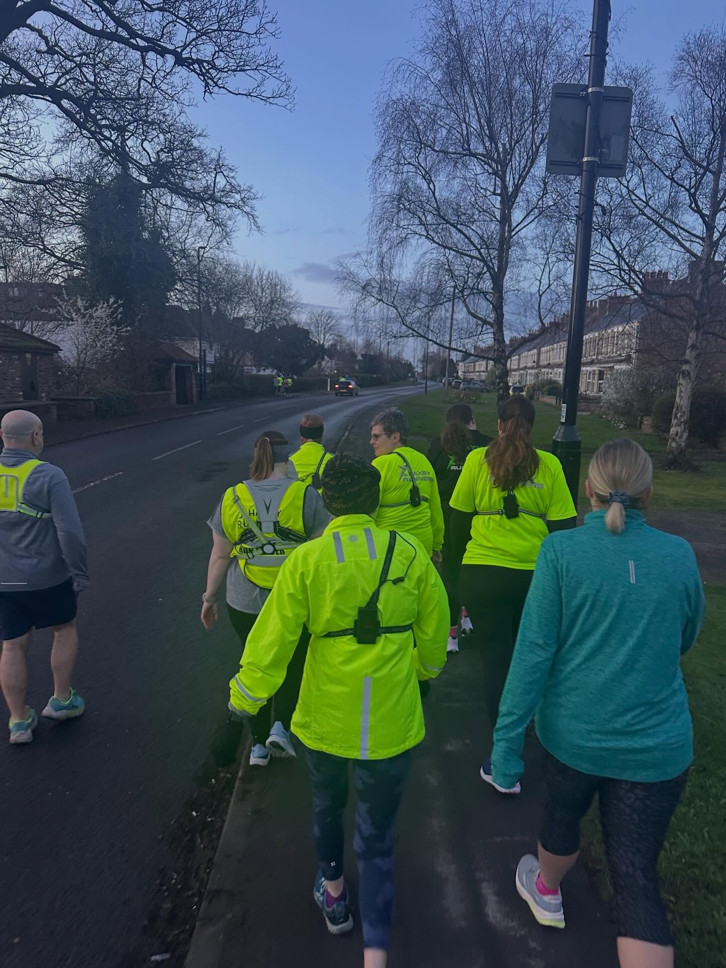 Group of people walking on a sidewalk in the evening, wearing yellow high-visibility vests, with a residential neighborhood and trees in the background.