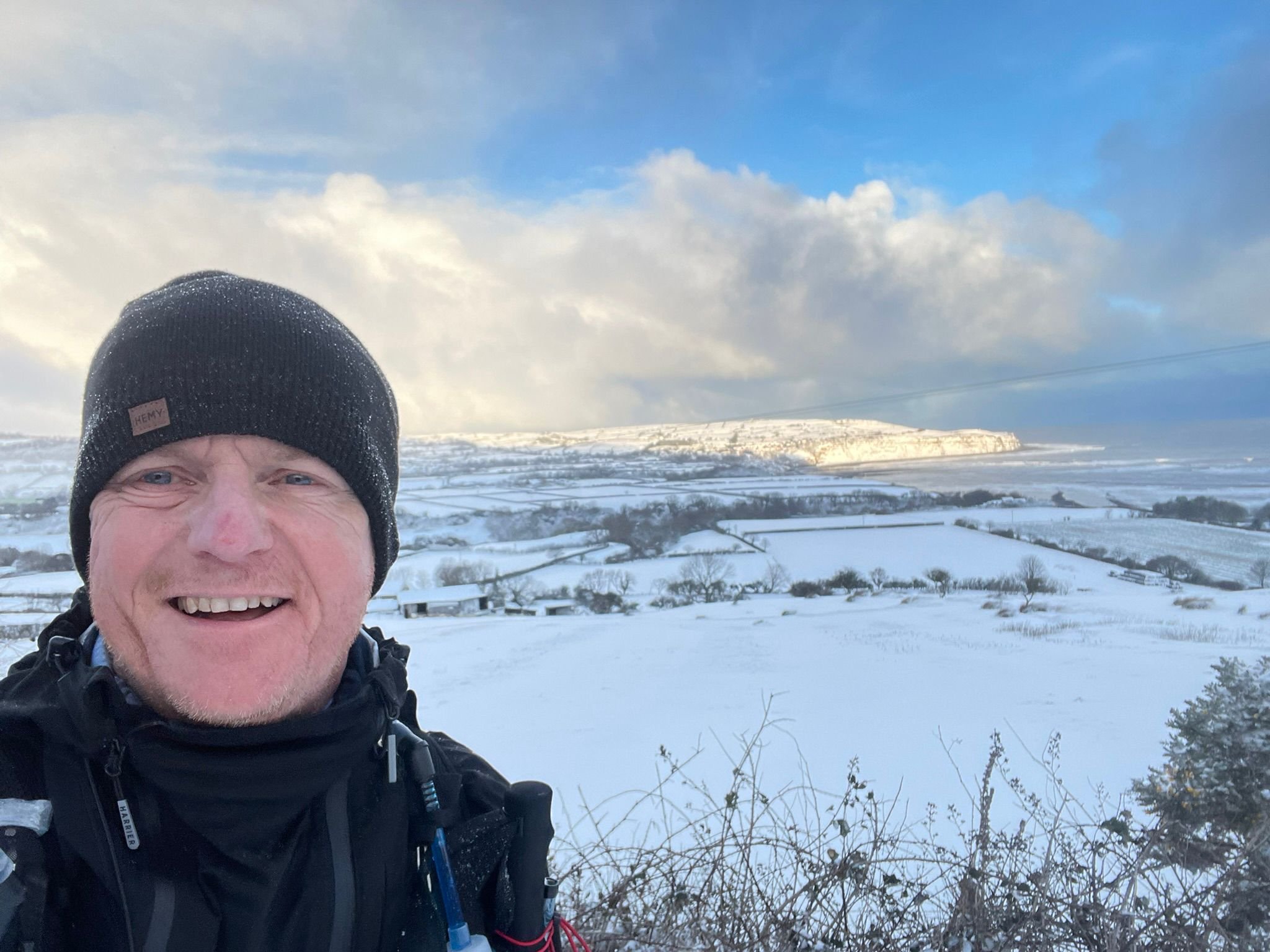 A man smiling outdoors on a snowy landscape under a partly cloudy sky, wearing a black beanie and a black jacket.