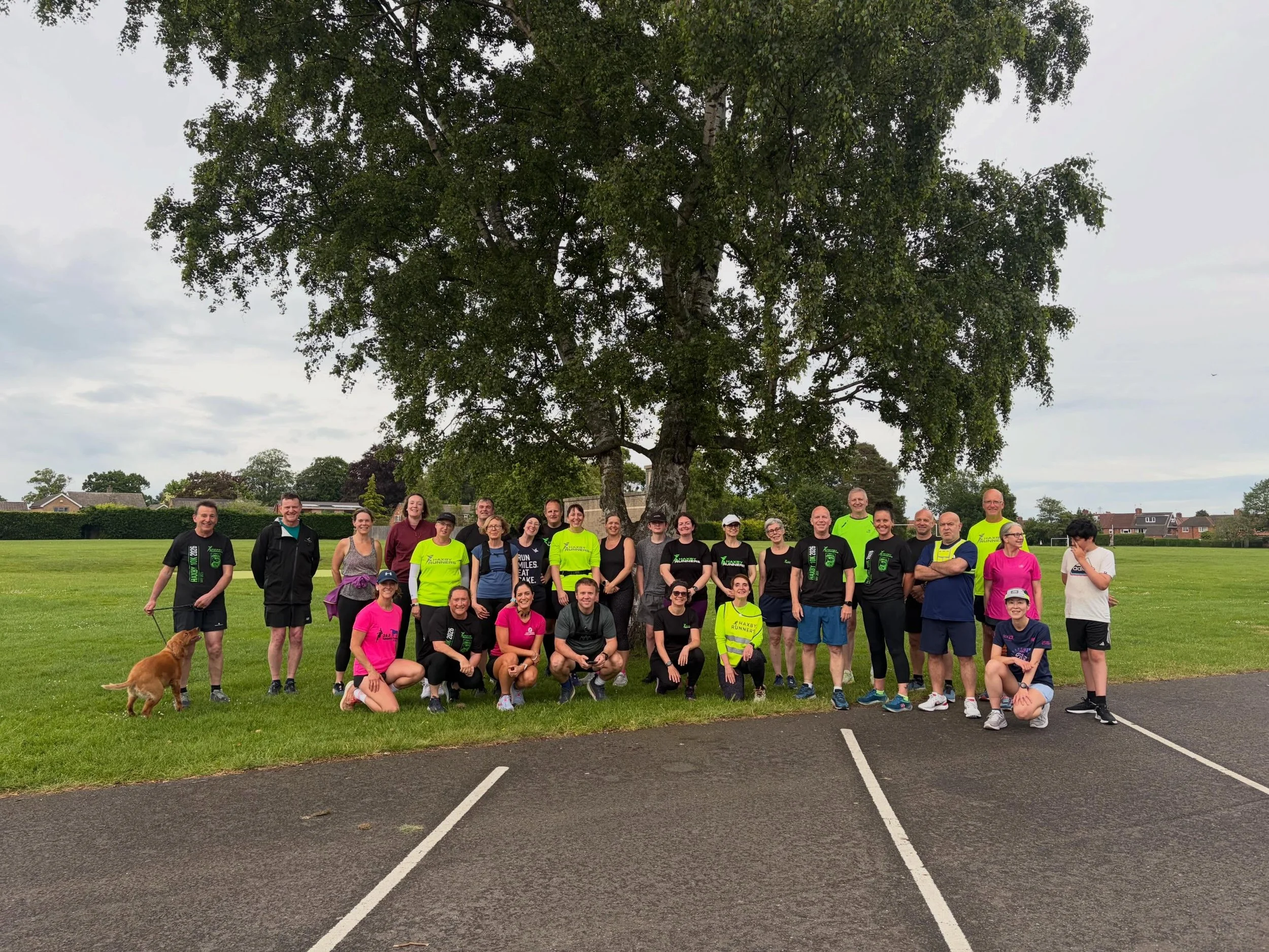 A group of people gathered outdoors on a grassy field with a large tree in the background. They are dressed in athletic clothing, some wearing bright yellow shirts, and are posing for a photo with smiles. There is a small brown dog in the foreground 