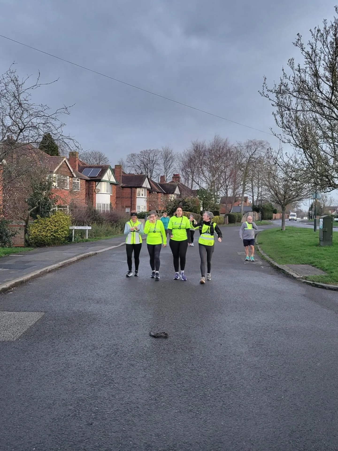A group of five women and one man walking on a suburban street, all wearing bright yellow running vests. The street is wet and lined with trees and houses under a cloudy sky.