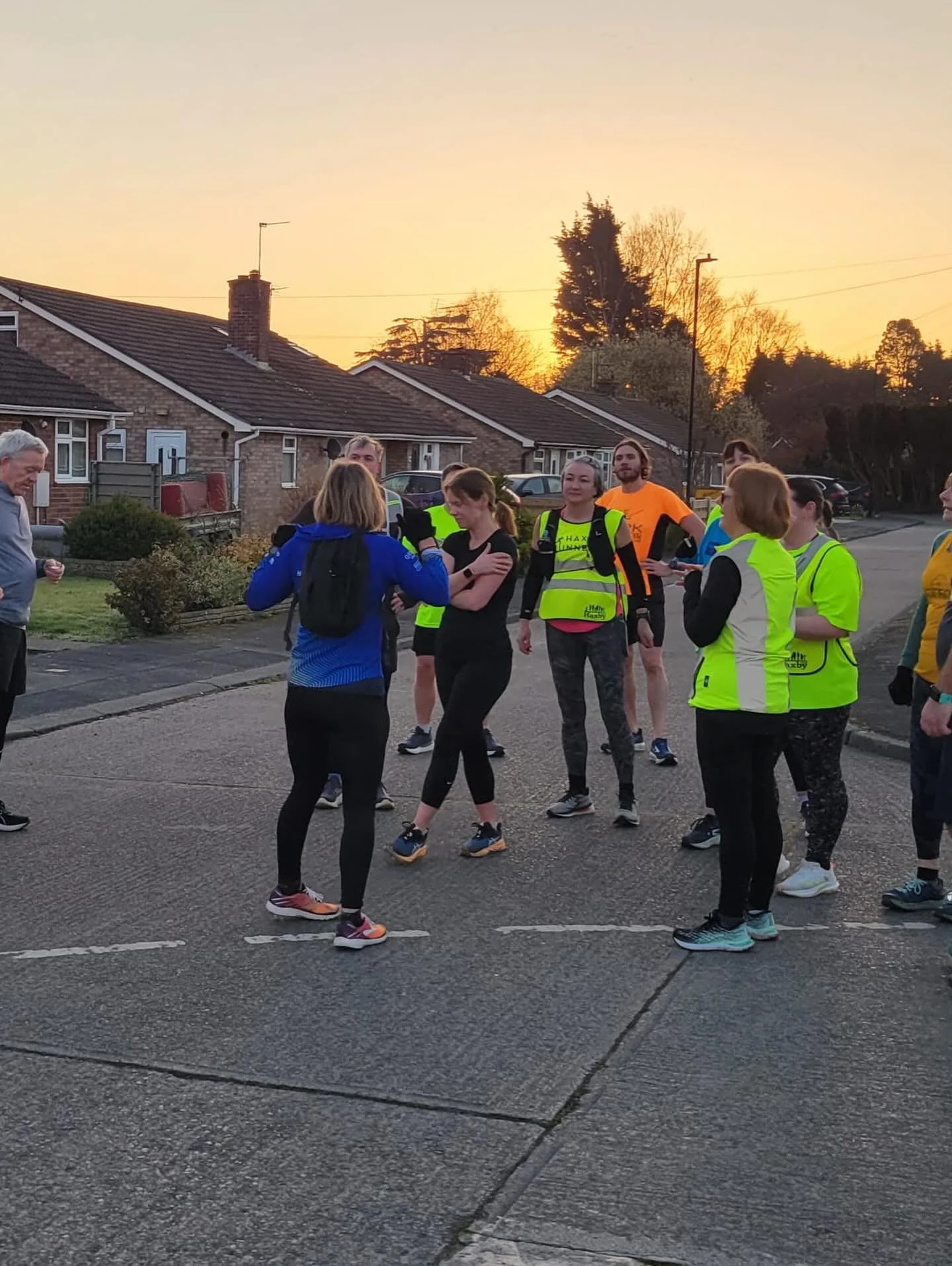 A group of runners in athletic clothing and vests gathered on a residential street at sunset, preparing for a run or race.