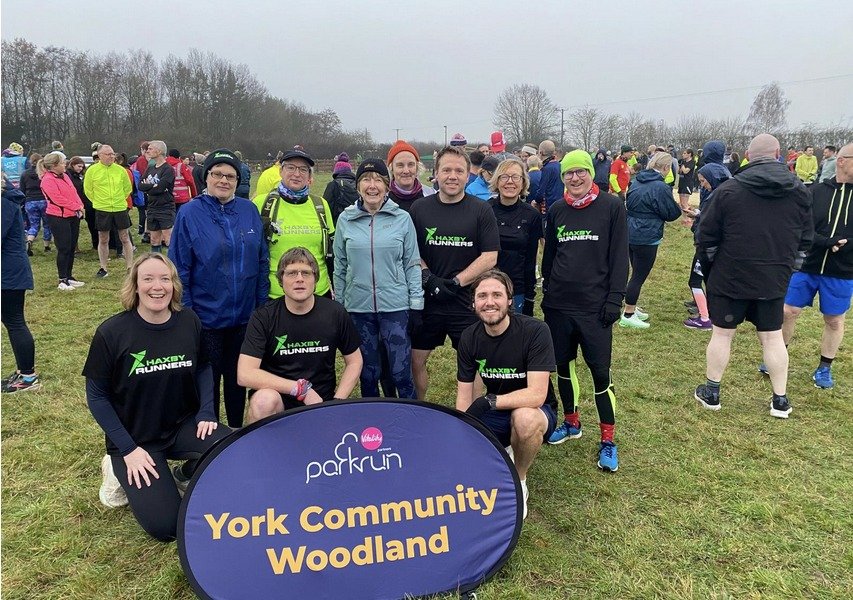 Group of people at a park run event in York Woodland, some wearing running gear with 'Haxby Runners' logo, standing behind a sign that reads 'York Community Woodland', on a grassy area with trees and other participants in the background.