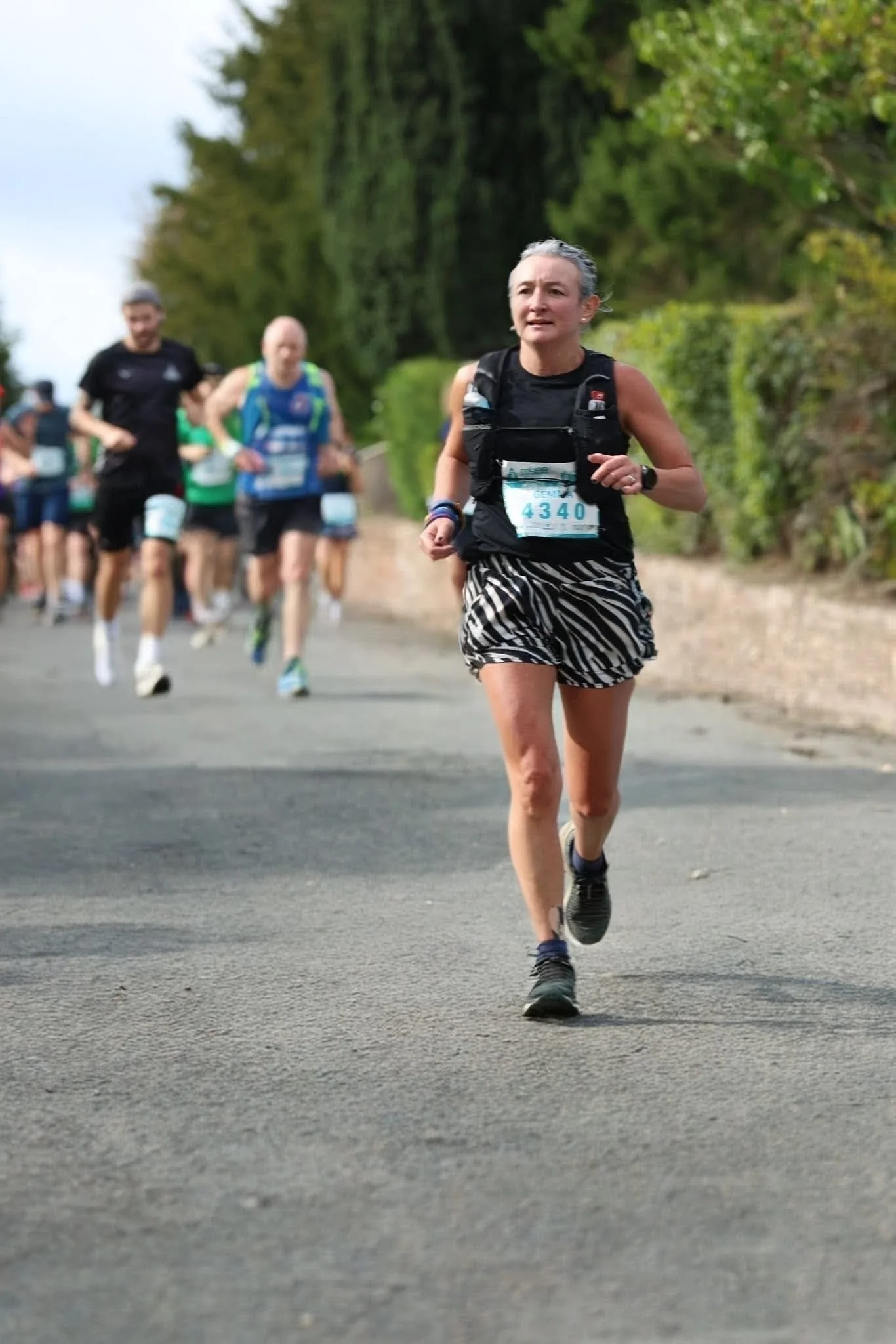 A group of runners participating in a race on a paved road surrounded by greenery. The woman in front is wearing a black vest, zebra-patterned shorts, and running shoes.