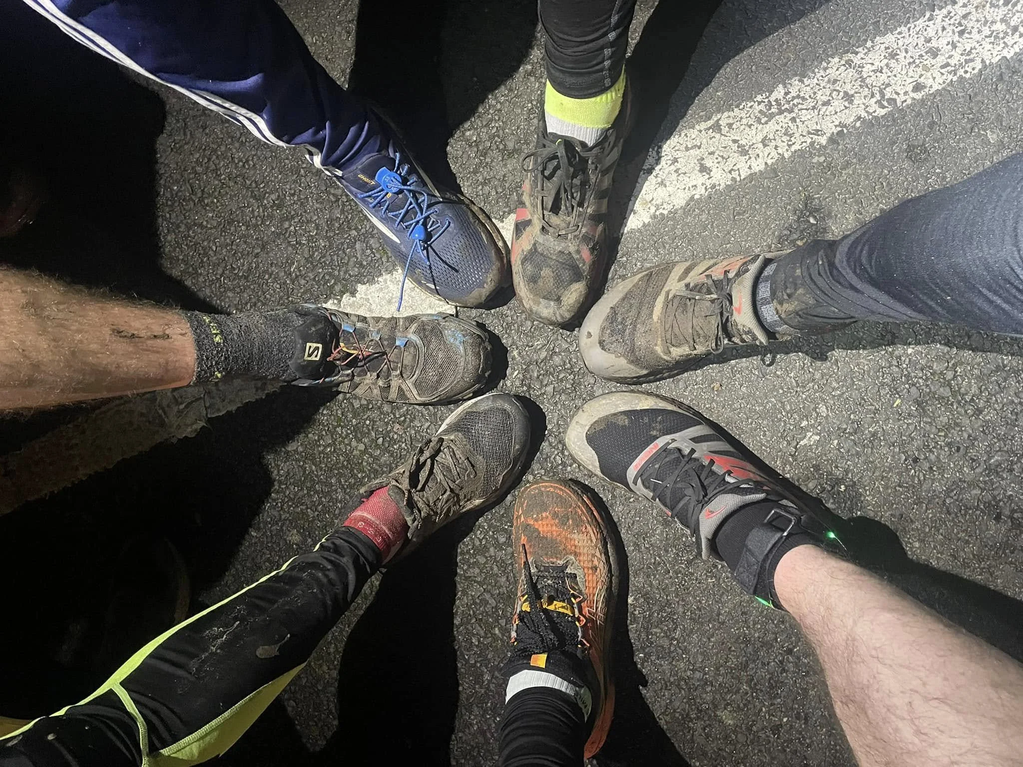 A group of runners' shoes arranged in a circle on a paved road, showing various athletic footwear, some dirty from trail running.