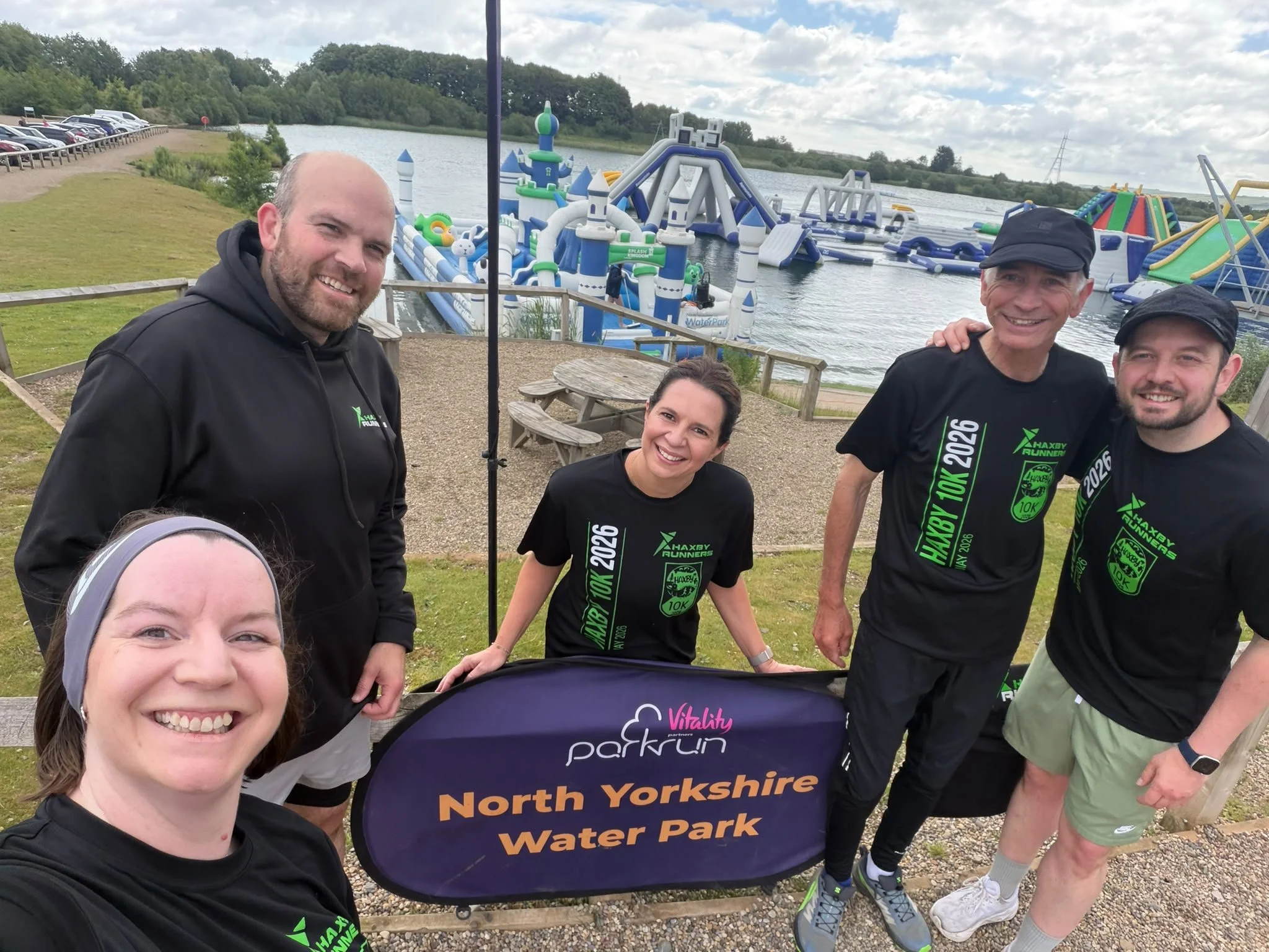 A group of five people smiling at the camera, standing outdoors near a lake with inflatables and water park equipment in the background, at North Yorkshire Water Park.