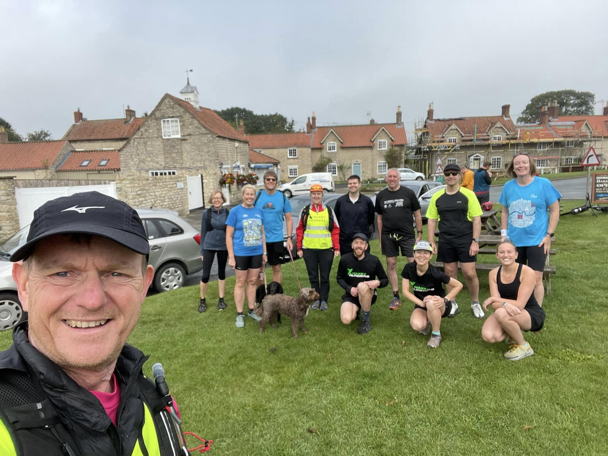 A group of people, including a man taking a selfie, gathered outdoors in front of houses, with some people holding leashes with dogs, in a grassy area on a cloudy day.