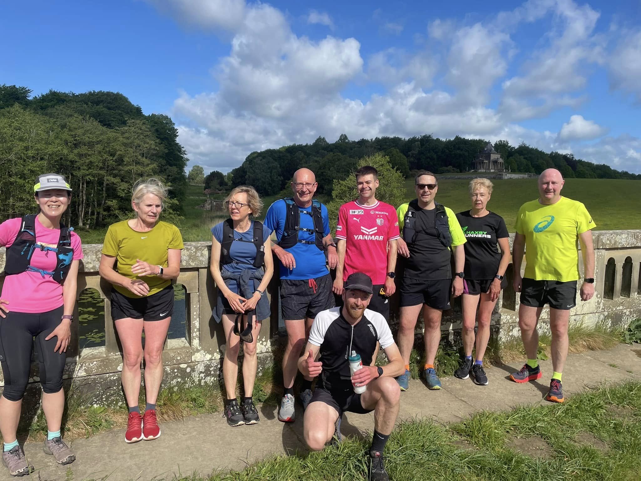 A group of nine runners posing outdoors on a bridge with greenery, hills, and a small building in the background.