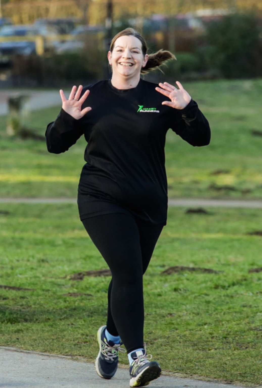 A woman running outdoors on a grassy path, smiling and waving, wearing a black long-sleeve shirt and black leggings.