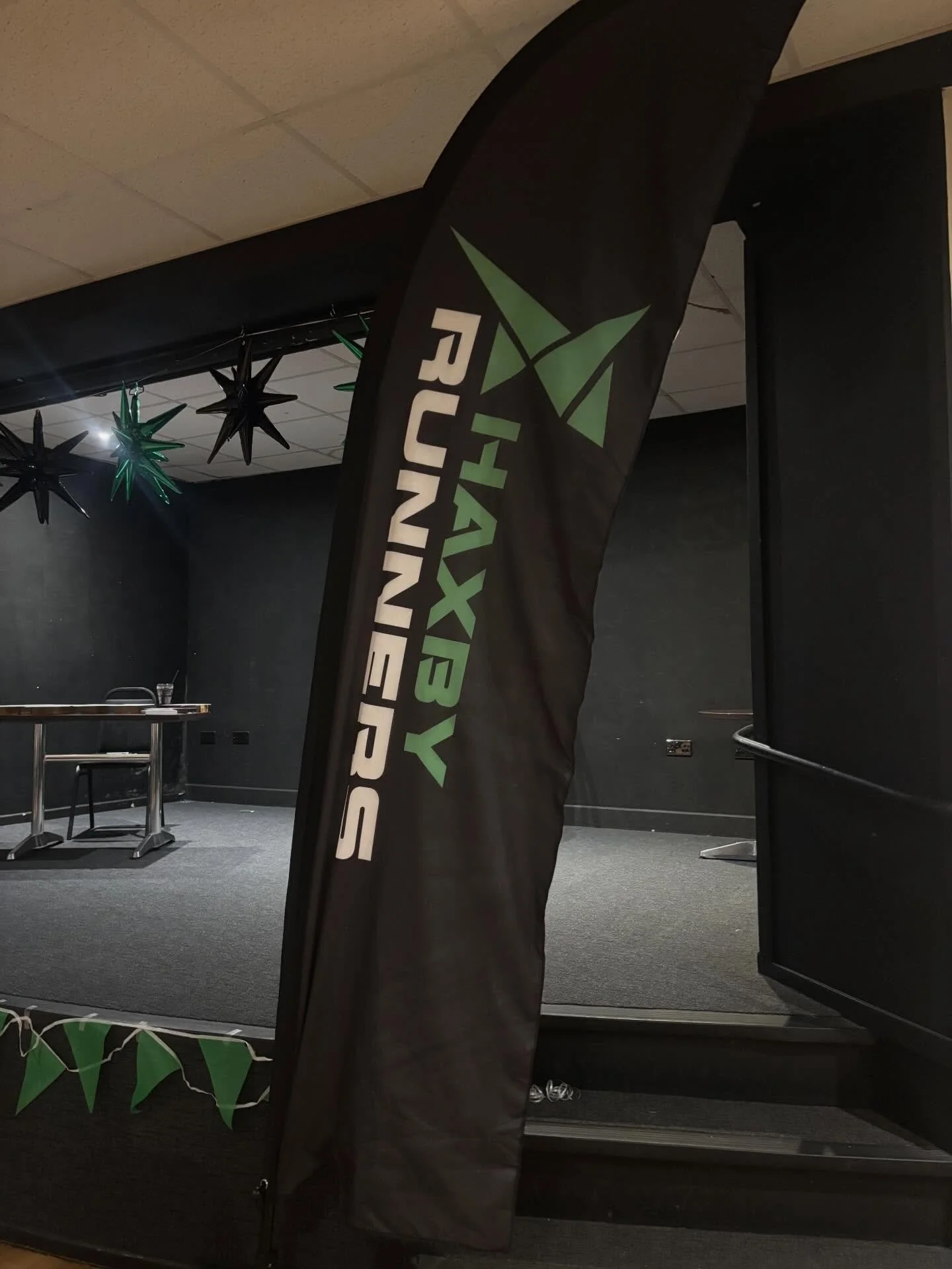 An indoor stage with black walls and ceiling, decorated with black and green star-shaped hanging decorations, and a black vertical flag with green and white text that reads 'HAXBY FUNNIESR' and features a green geometric design.