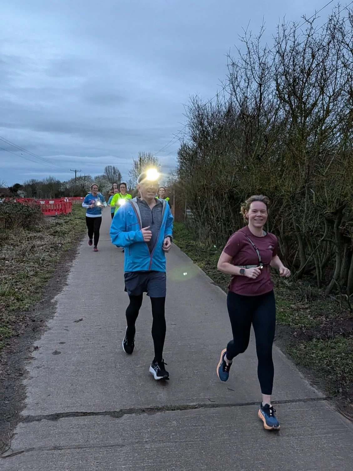 Group of people jogging on a paved path outdoors, some wearing headlamps or running gear, during dusk or early evening.