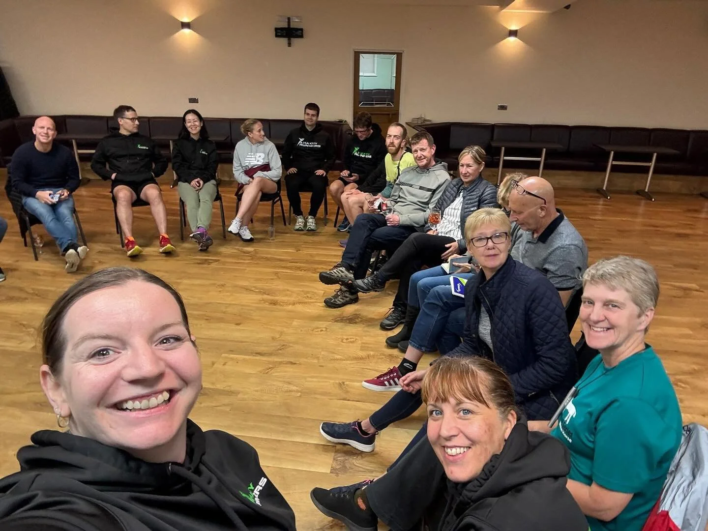 People sitting in a large circle inside a room with wooden floors, smiling and taking selfies.
