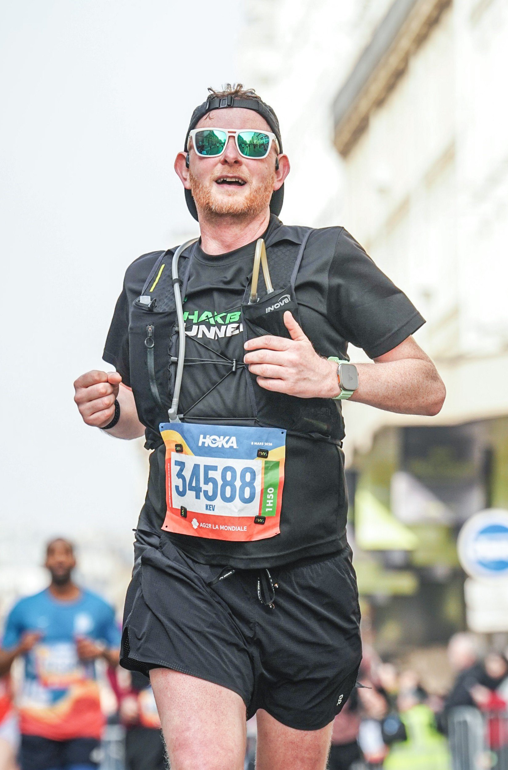 Male marathon runner in black athletic attire, sunglasses, and a hydration vest, smiling as he runs in a race.