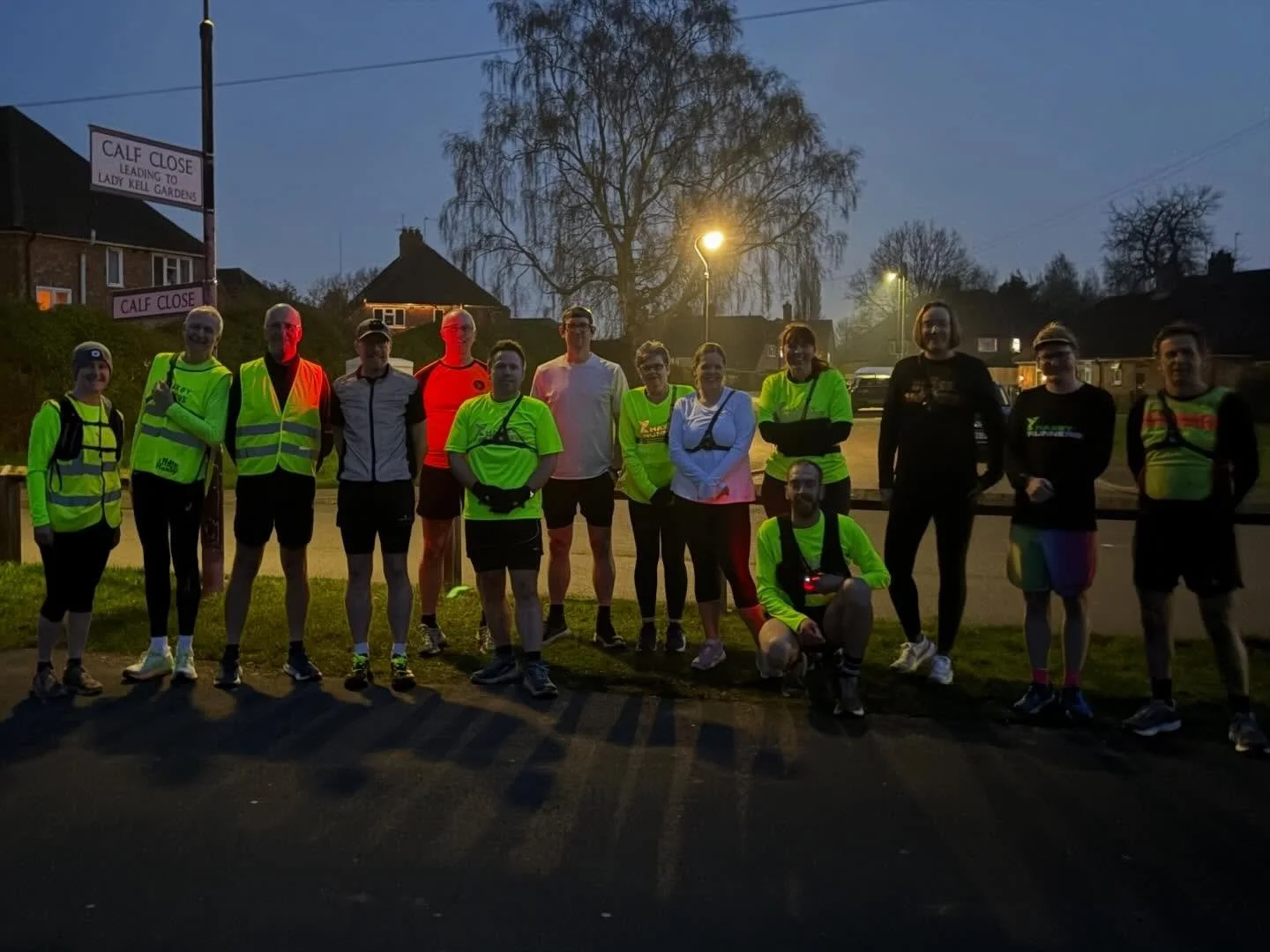 Group of runners in bright athletic clothing and reflective vests standing on a sidewalk at dusk, with streetlights and houses in the background.