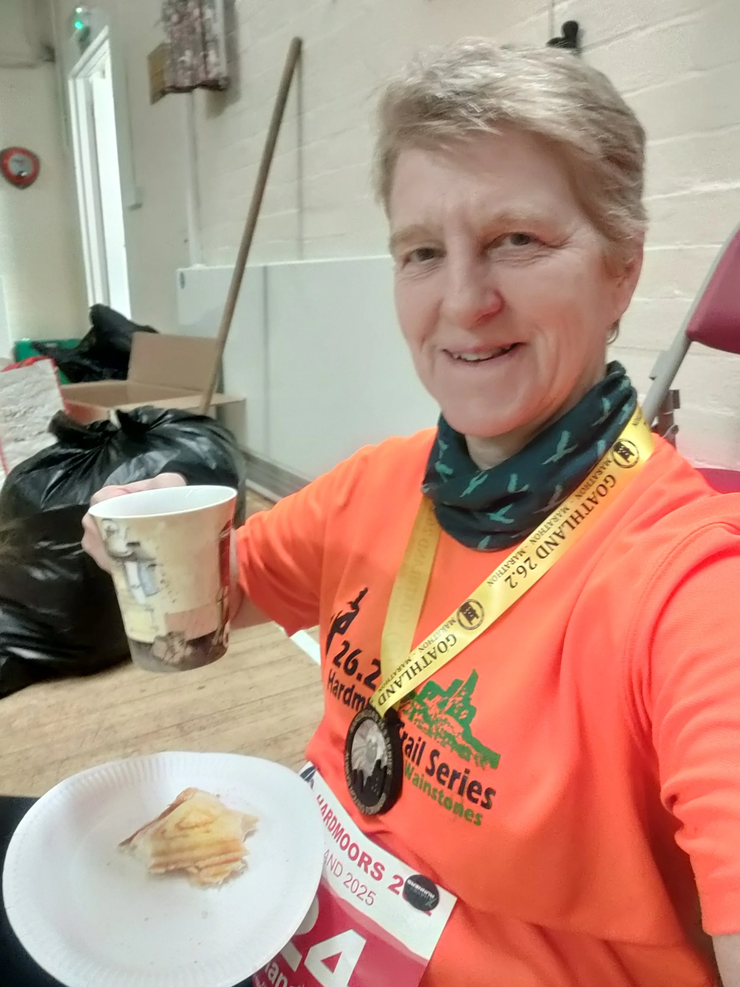 A smiling woman wearing an orange T-shirt, a medal around her neck, and a trail running bib, holding a paper plate with a slice of cake and a coffee cup, sitting indoors with garbage bags and cleaning tools in the background.