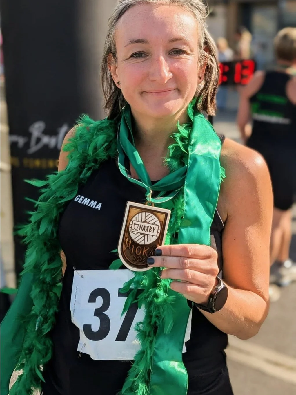 A smiling woman wearing black athletic clothing with the name 'GEMMA' on it, and a race bib number 37. She is holding a medal that reads '10K 2025' and a green feathered boa around her shoulders. She is outdoors, celebrating after completing a race.