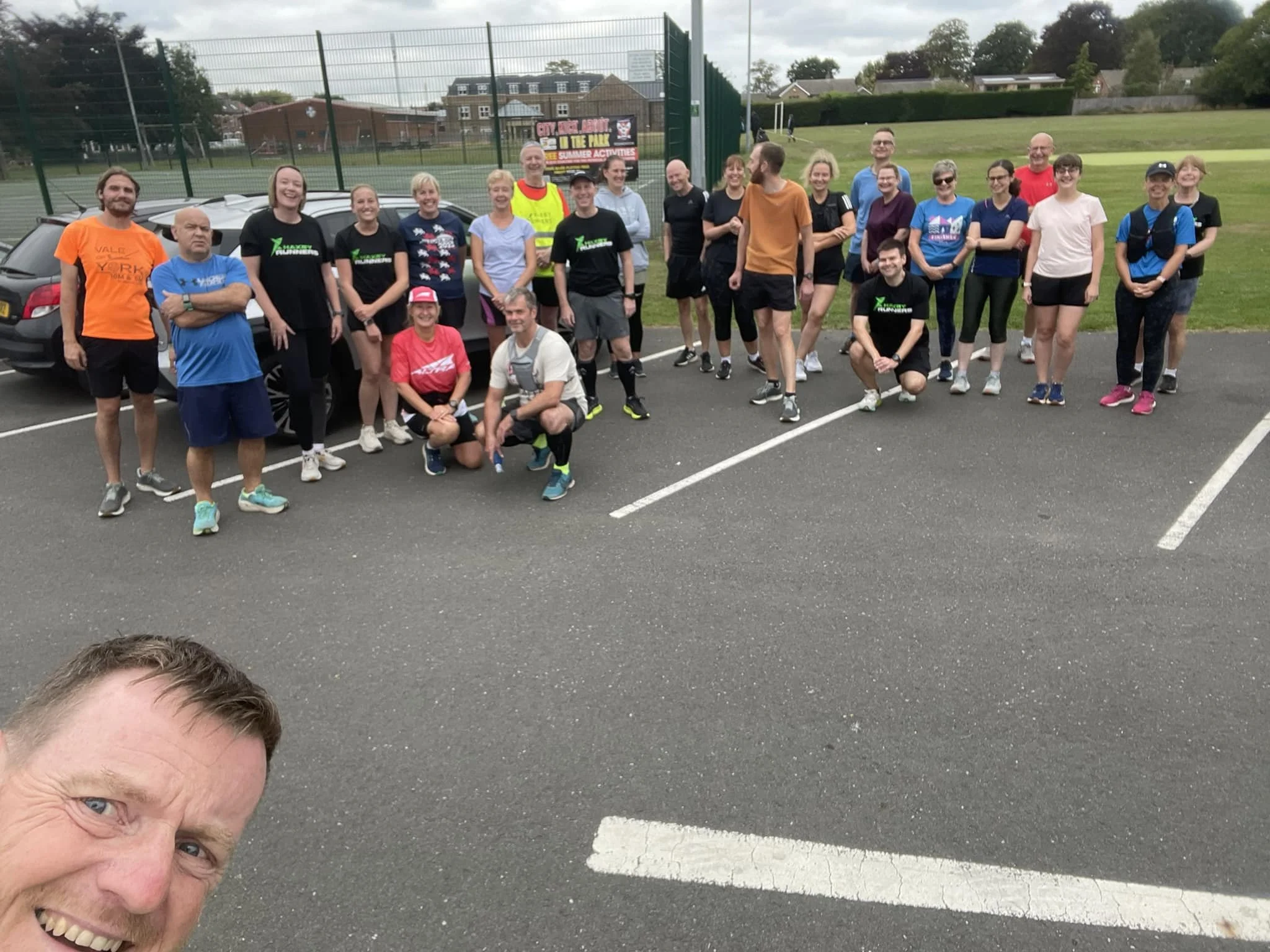 Group of people in athletic clothing standing and sitting in a parking lot near a sports field, posing for a photo, with a man taking a selfie in the foreground.