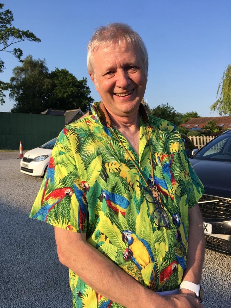 A smiling older man wearing a colorful Hawaiian shirt with parrots and tropical leaves, standing outdoors in a parking lot on a sunny day.