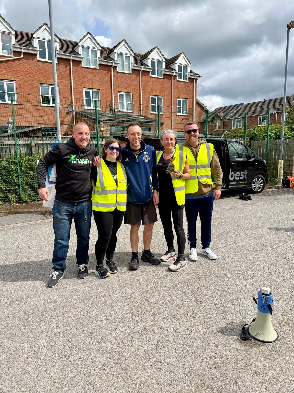 Five people standing together outdoors, smiling, four wearing high-visibility safety vests. Two women and two men are holding a small trophy or award. A black van with the word 'best' on it is parked behind them, with a row of brick houses and cloudy sky in the background.