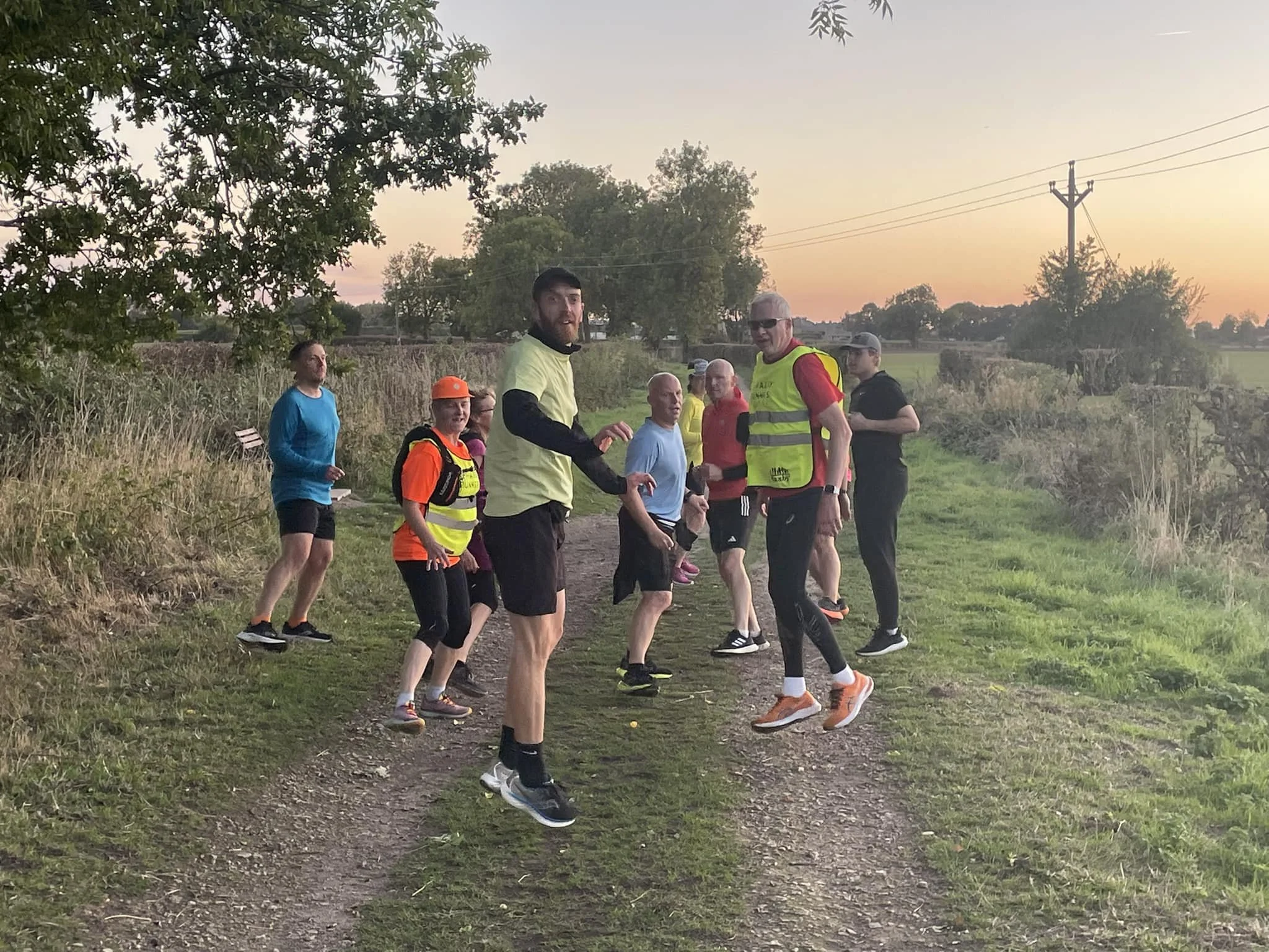 A group of runners stopping on a rural trail at sunset, some wearing bright athletic clothing and backpacks, with trees, power lines, and a sunset sky in the background.