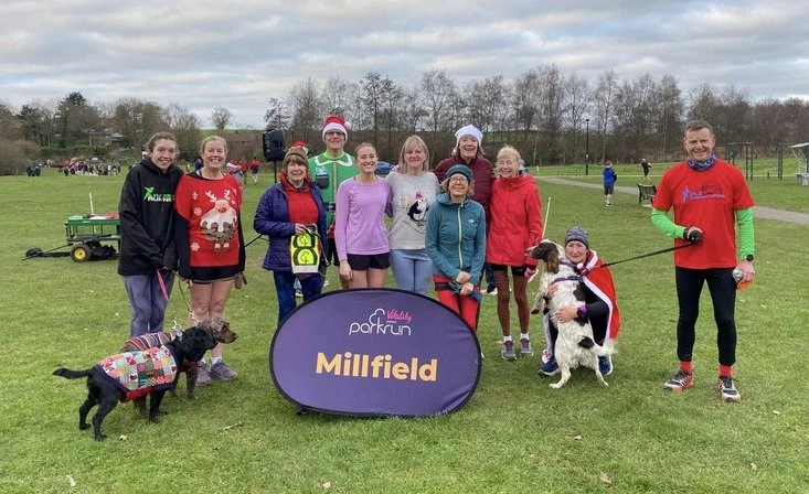 Group of people standing on a grassy field, many wearing holiday-themed clothing, with dogs and a Millfield parkrun sign in front, suggesting a festive outdoor event.