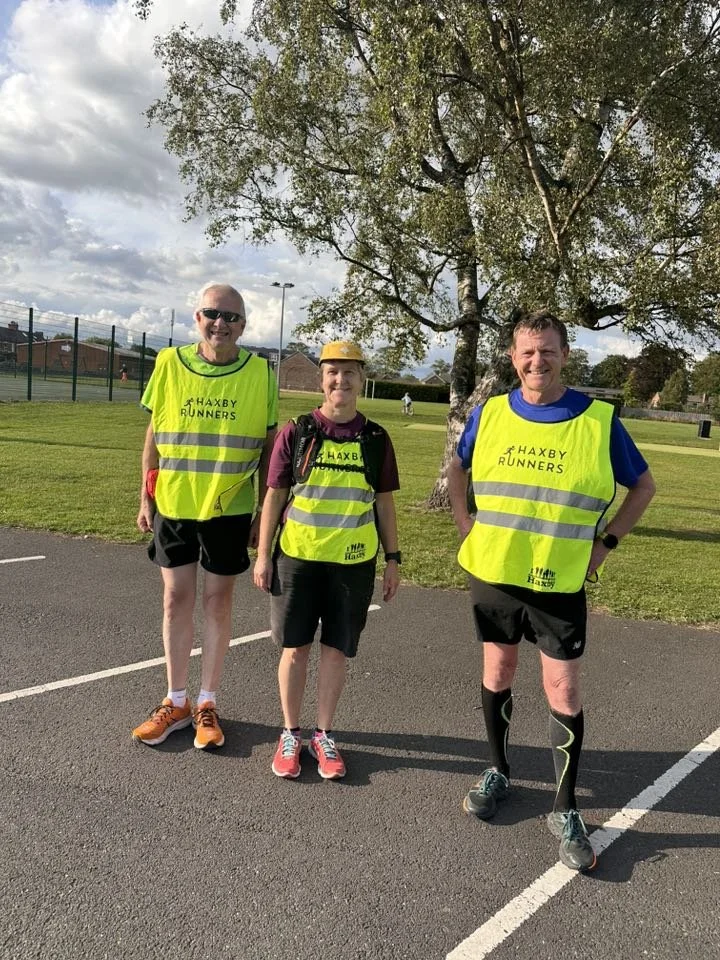 Three runners wearing yellow reflective vests with 'HAXBY RUNNERS' printed on them, standing on a paved path next to a grass field and a large tree, with a partly cloudy sky overhead.