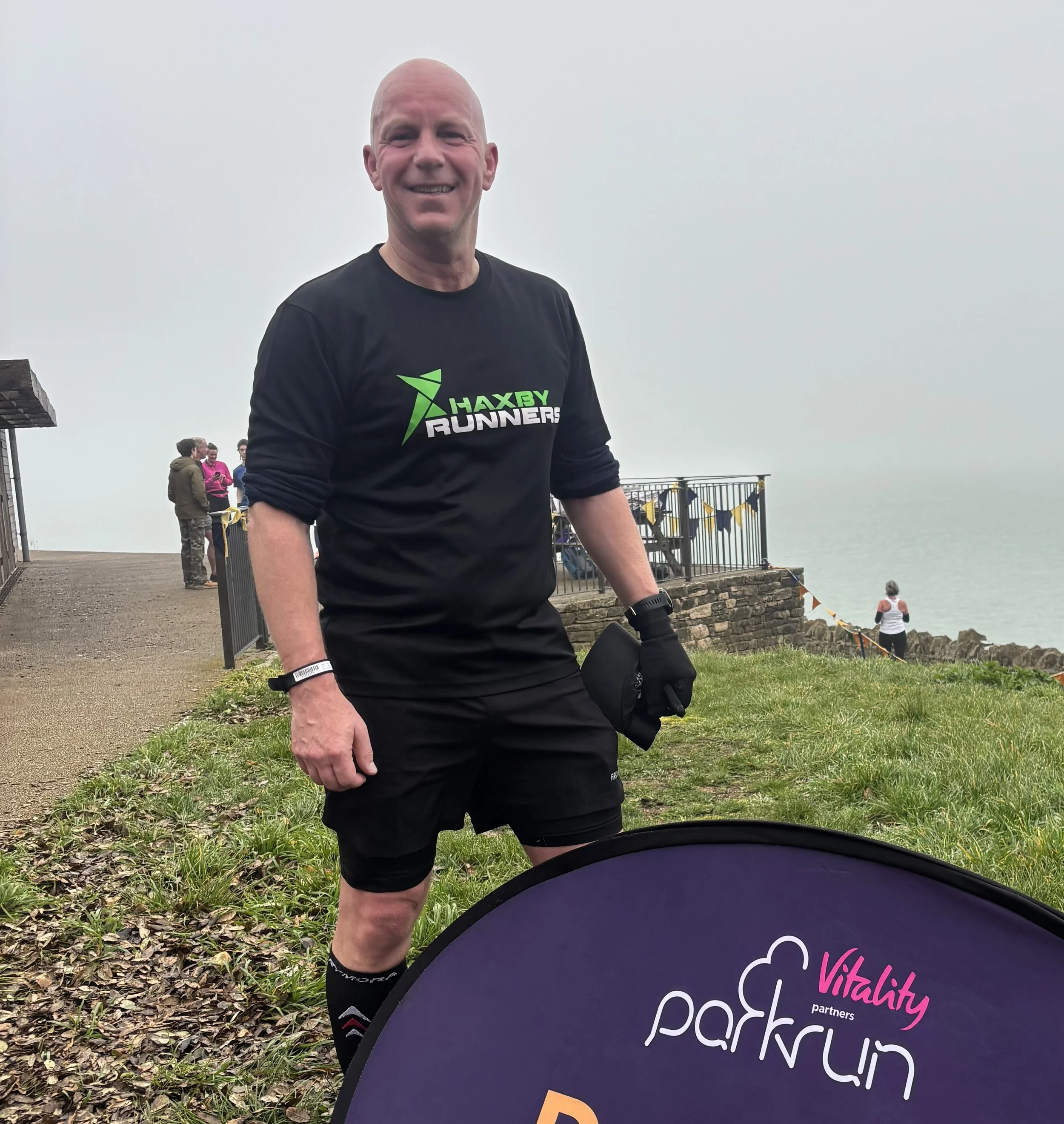 A smiling man wearing a black Haxby Runners shirt and black shorts, standing outdoors near a grassy area with a railing and a foggy body of water in the background, holding a black pouch, with a park run sign in front of him.