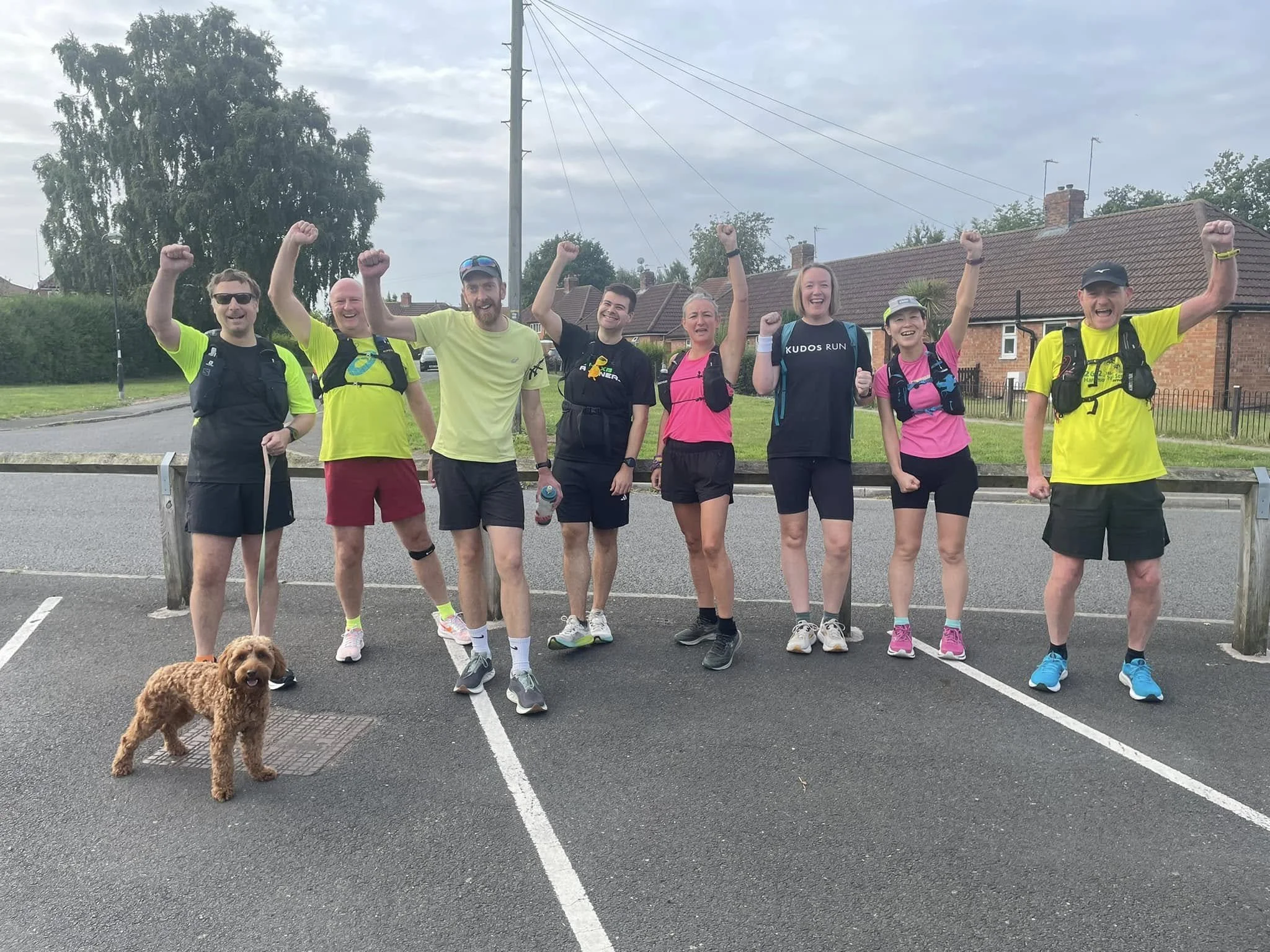 Group of eight people standing in a parking lot, celebrating with raised arms, dressed in athletic running gear, with a small dog in the foreground.