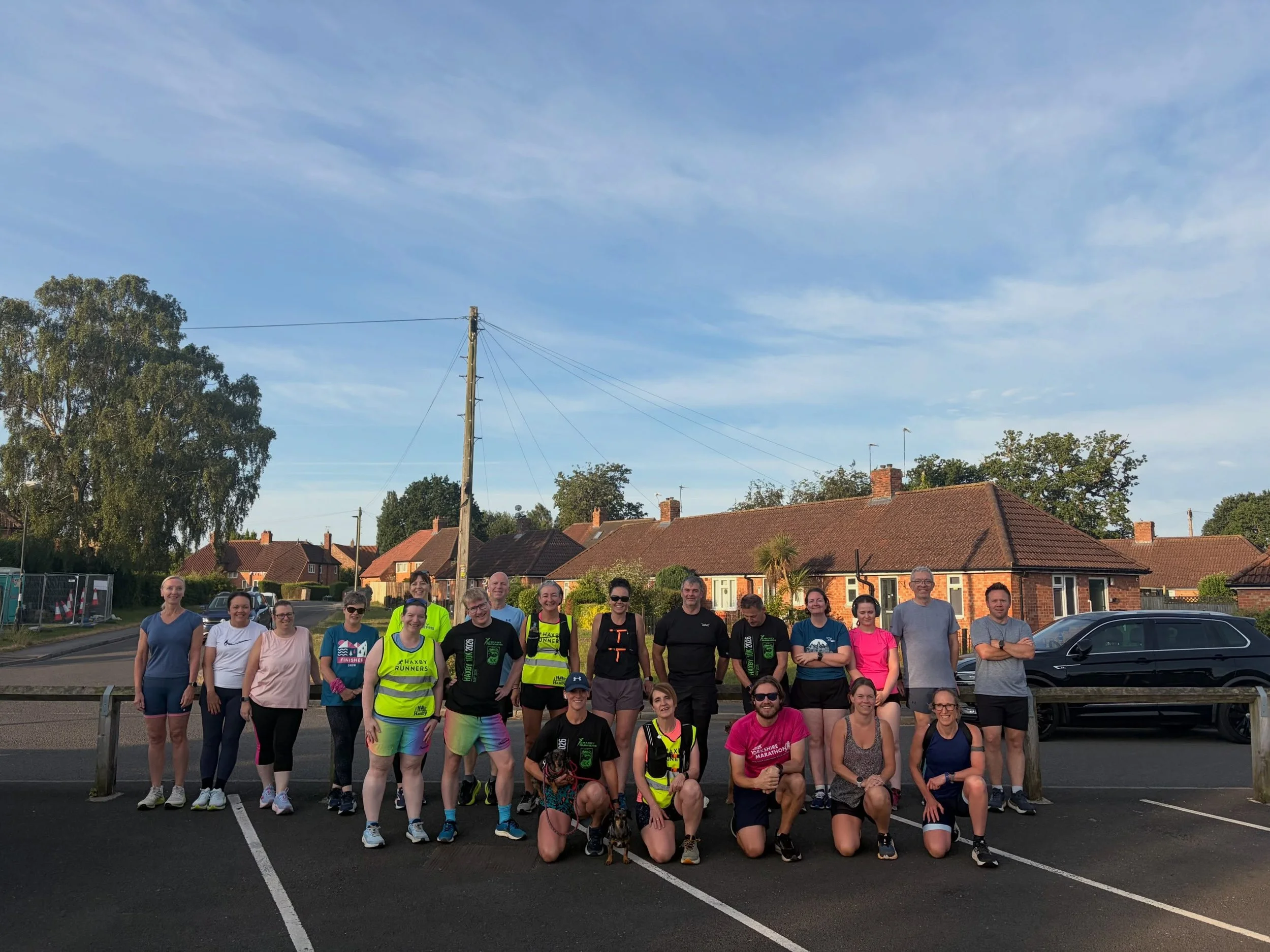 A group of people gathered outdoors in a parking lot, with some wearing running apparel and race medals, likely after participating in a running event, with houses, trees, and a blue sky in the background.