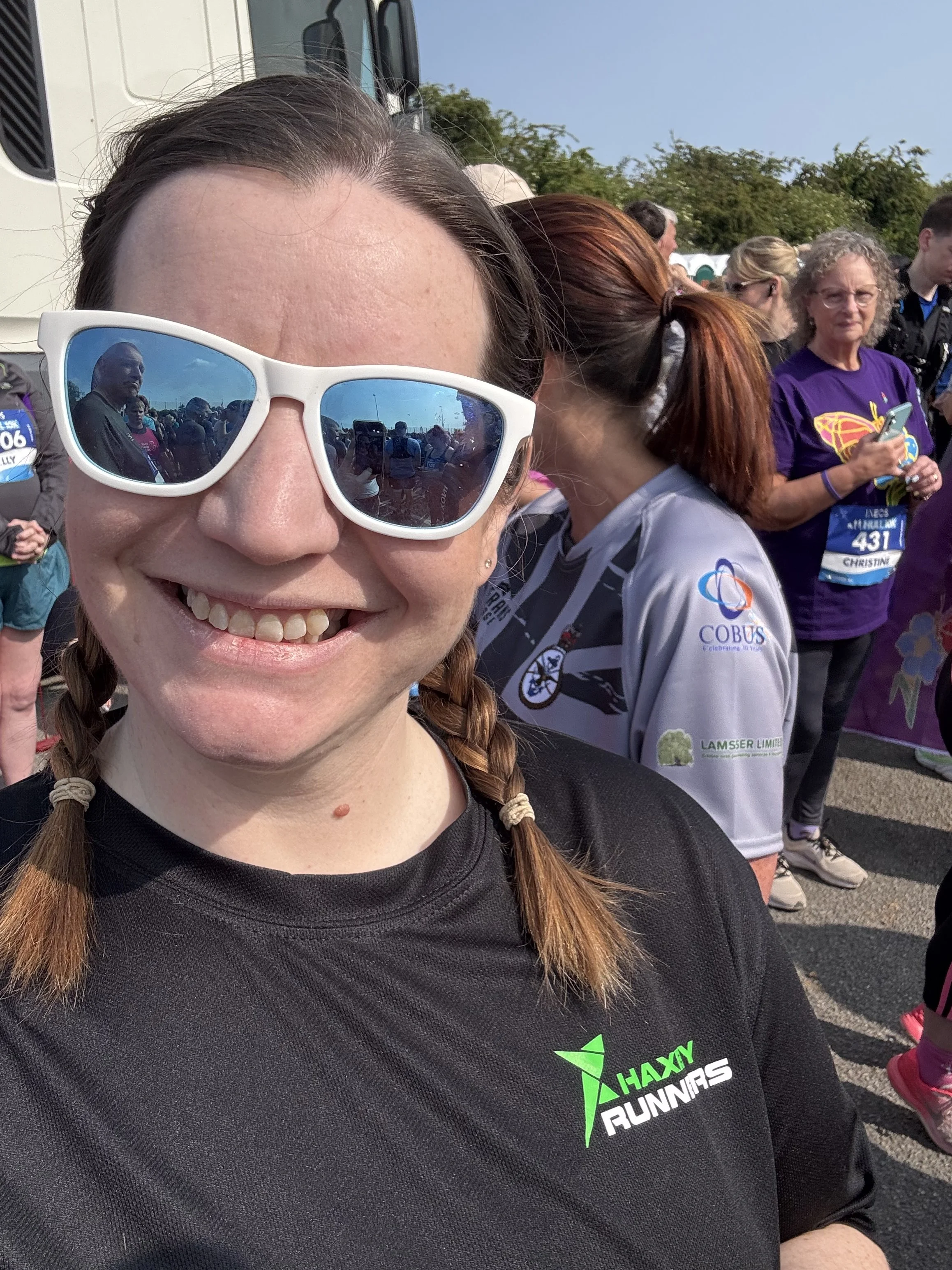 A woman wearing sunglasses and a black T-shirt with green and white text that says 'Haxby Runners,' smiling at the camera at a race event with other runners in the background.