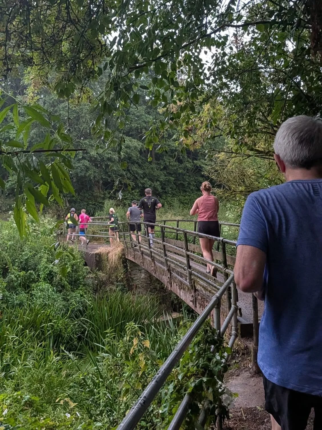 A group of people running and walking on a small wooden bridge in a lush green park with dense foliage.