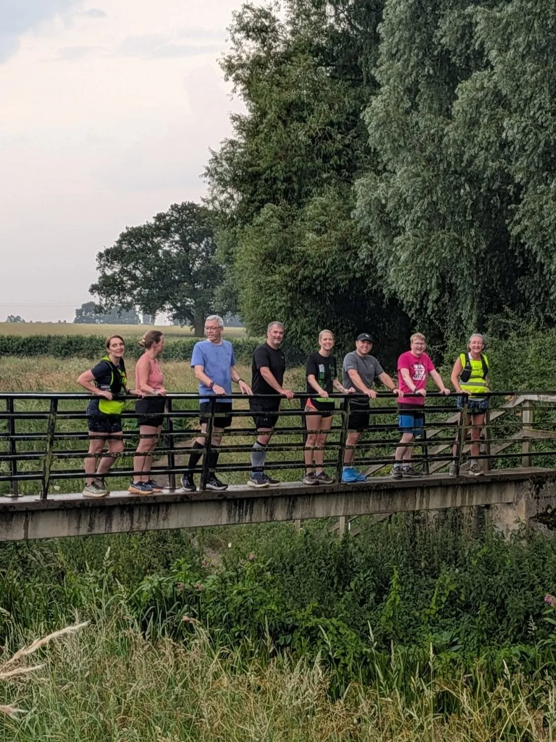 A group of eight people standing on a small bridge in a green rural area, surrounded by trees and fields.