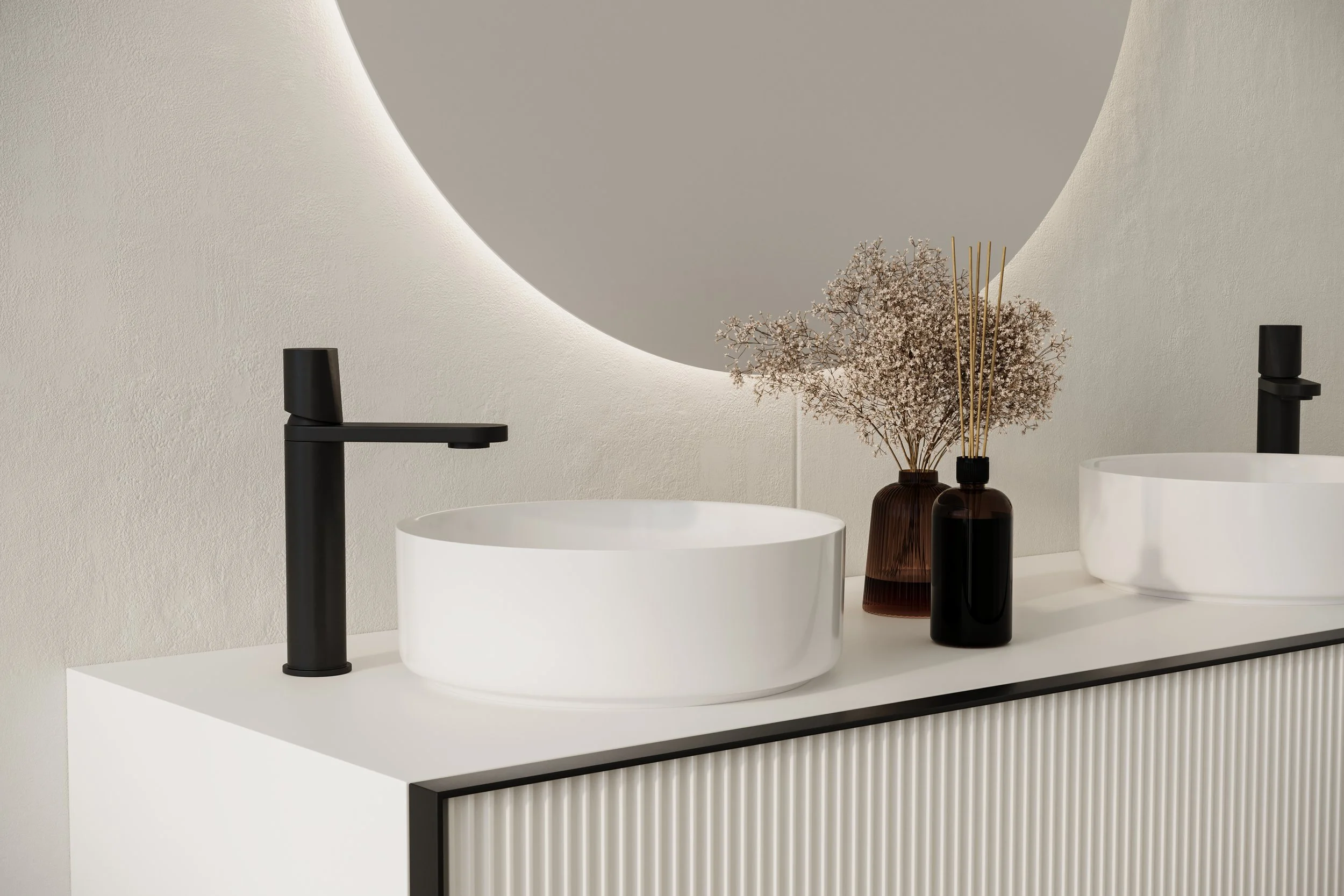 A modern bathroom sink with a white vessel sink, a black faucet, and decorative vases, including one with dried flowers, against a minimalist wall with a curved design and ambient lighting.