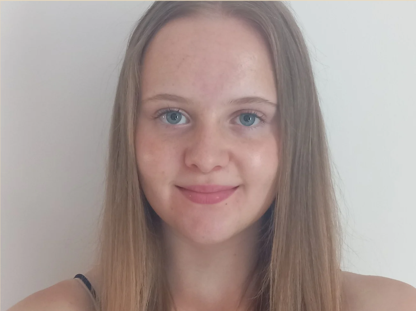 Portrait of a young woman with long, straight, blonde hair and blue eyes, smiling softly against a plain, light background.
