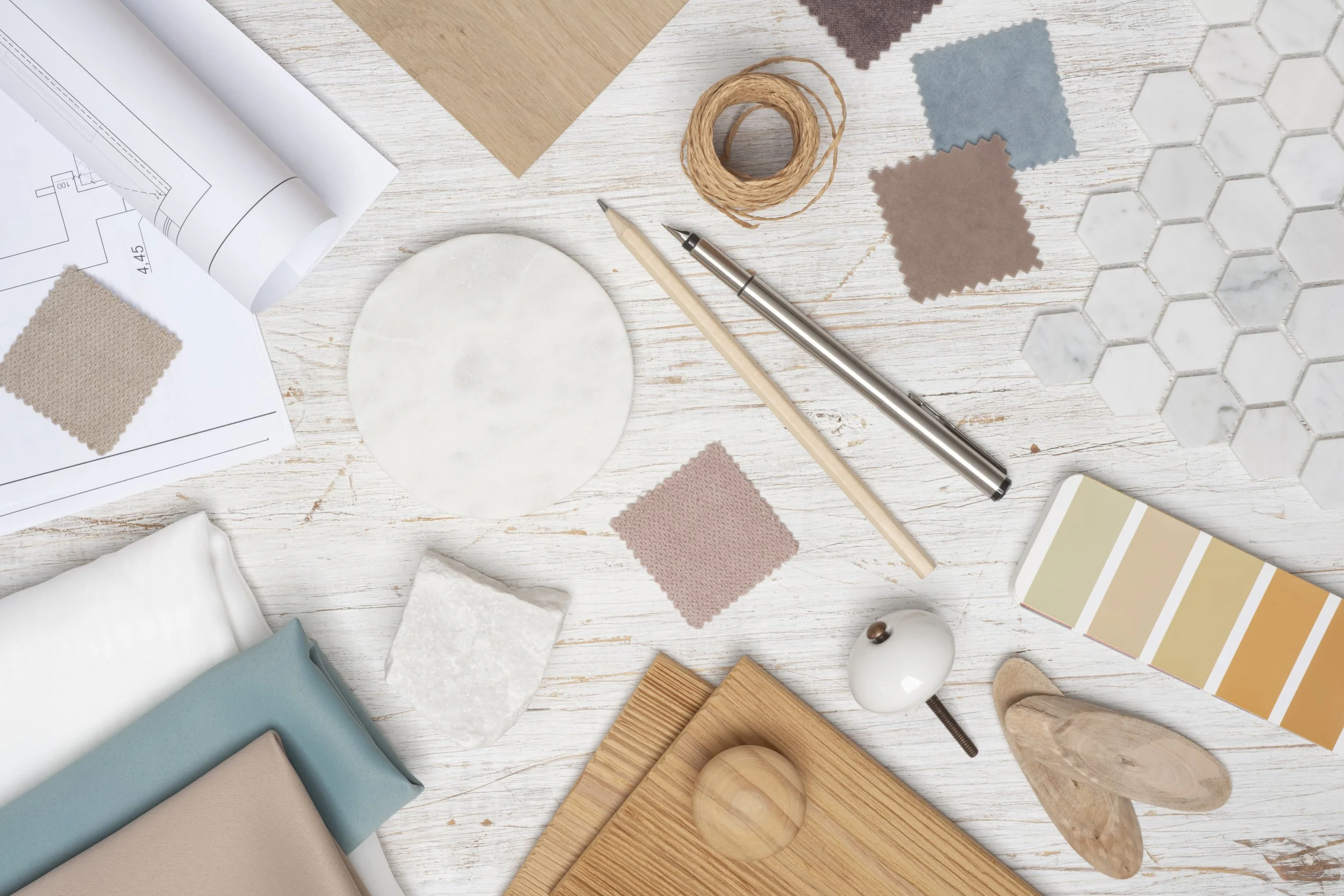 A flat lay of interior design materials including fabric swatches, paint colour samples, wooden samples, marble pattern, interior plans, and decorative objects on a white wooden surface.
