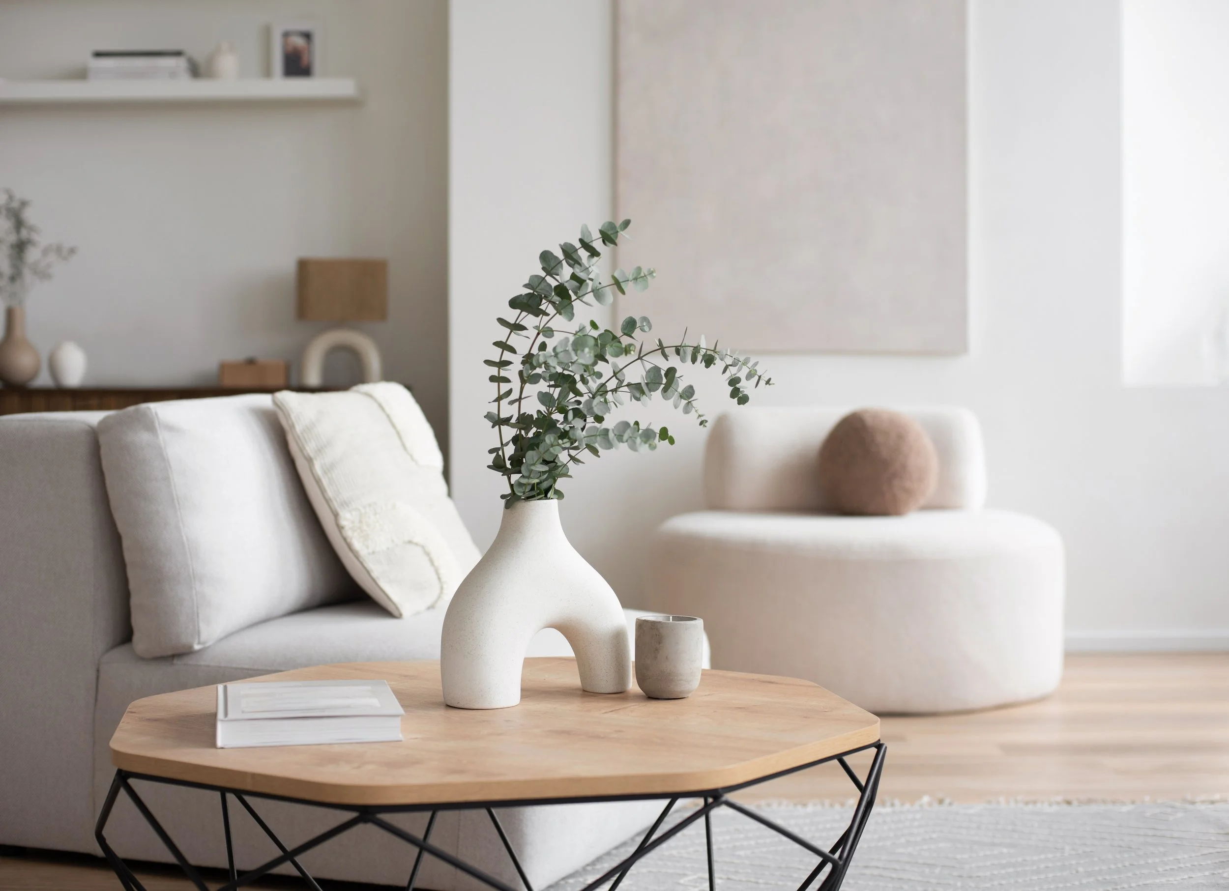 Living room with a light-colored sofa, beige cushions, wooden coffee table with a white book and ceramic vases, background wall with shelf, artwork, and decorative objects, modern and minimalist design.