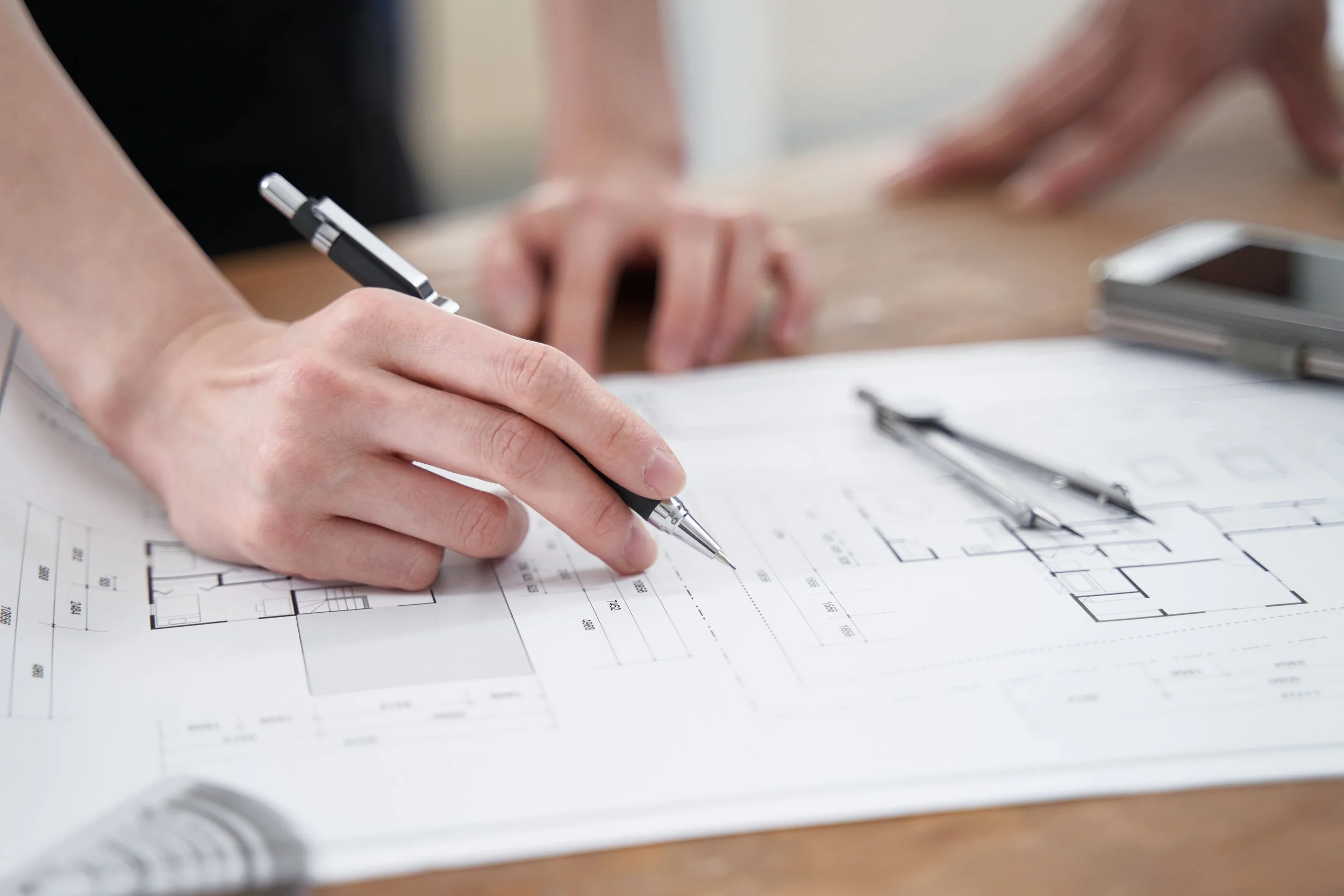 Close-up of a person's hand holding a pen and marking on an technical floor plan, with a pen placed on the blueprint in the background, on a wooden table.