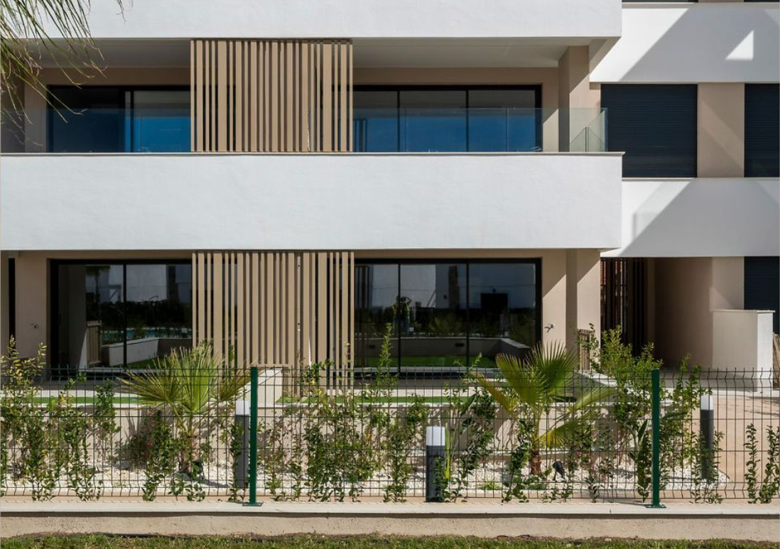 Edificio moderno de varios pisos con balcones y ventanas grandes, rodeado por una cerca metálica y plantas ornamentales en el jardín delantero.