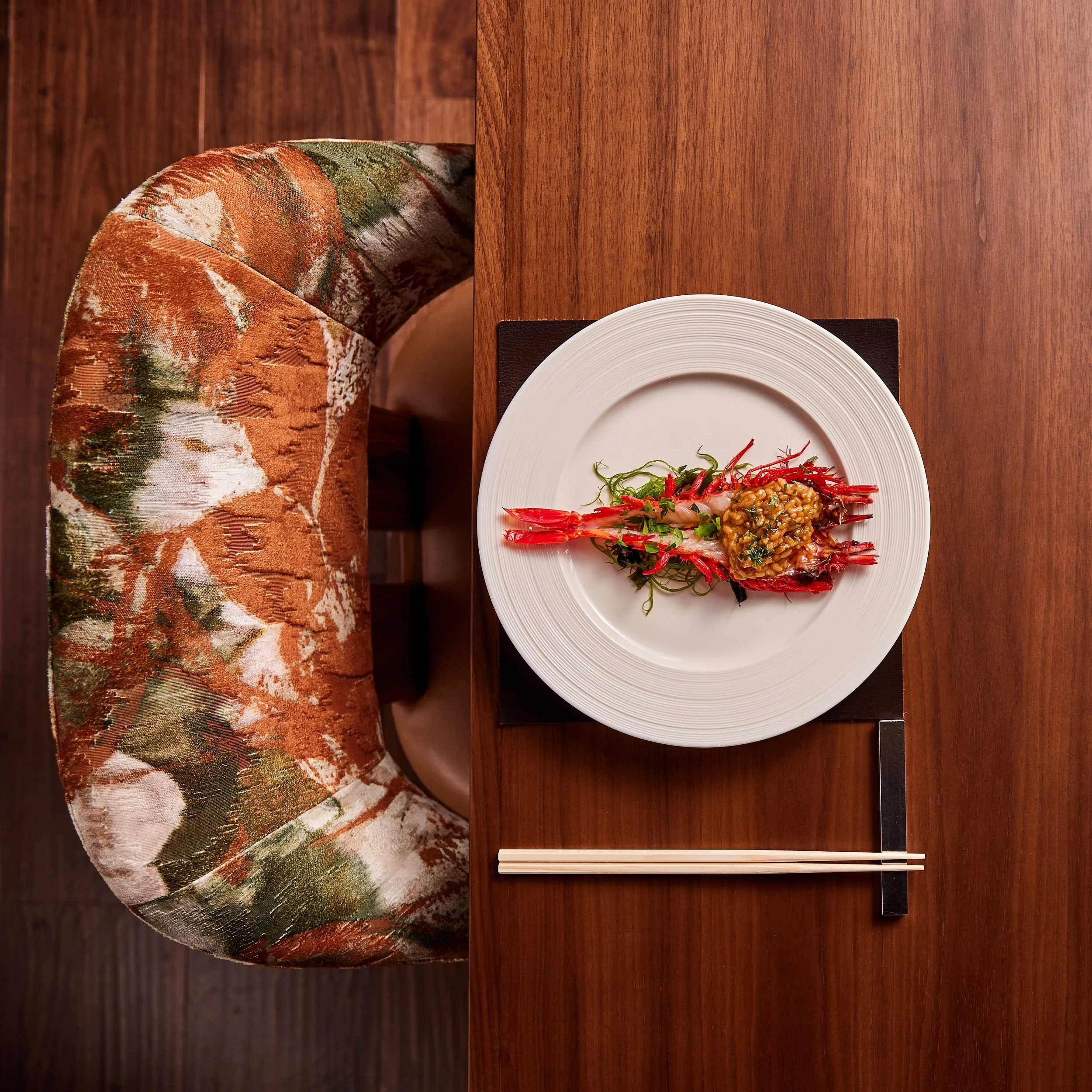 A wooden dining table with a white plate, a pair of chopsticks, and a dish of seafood and rice. Part of a patterned upholstered chair is visible on the left side of the image.