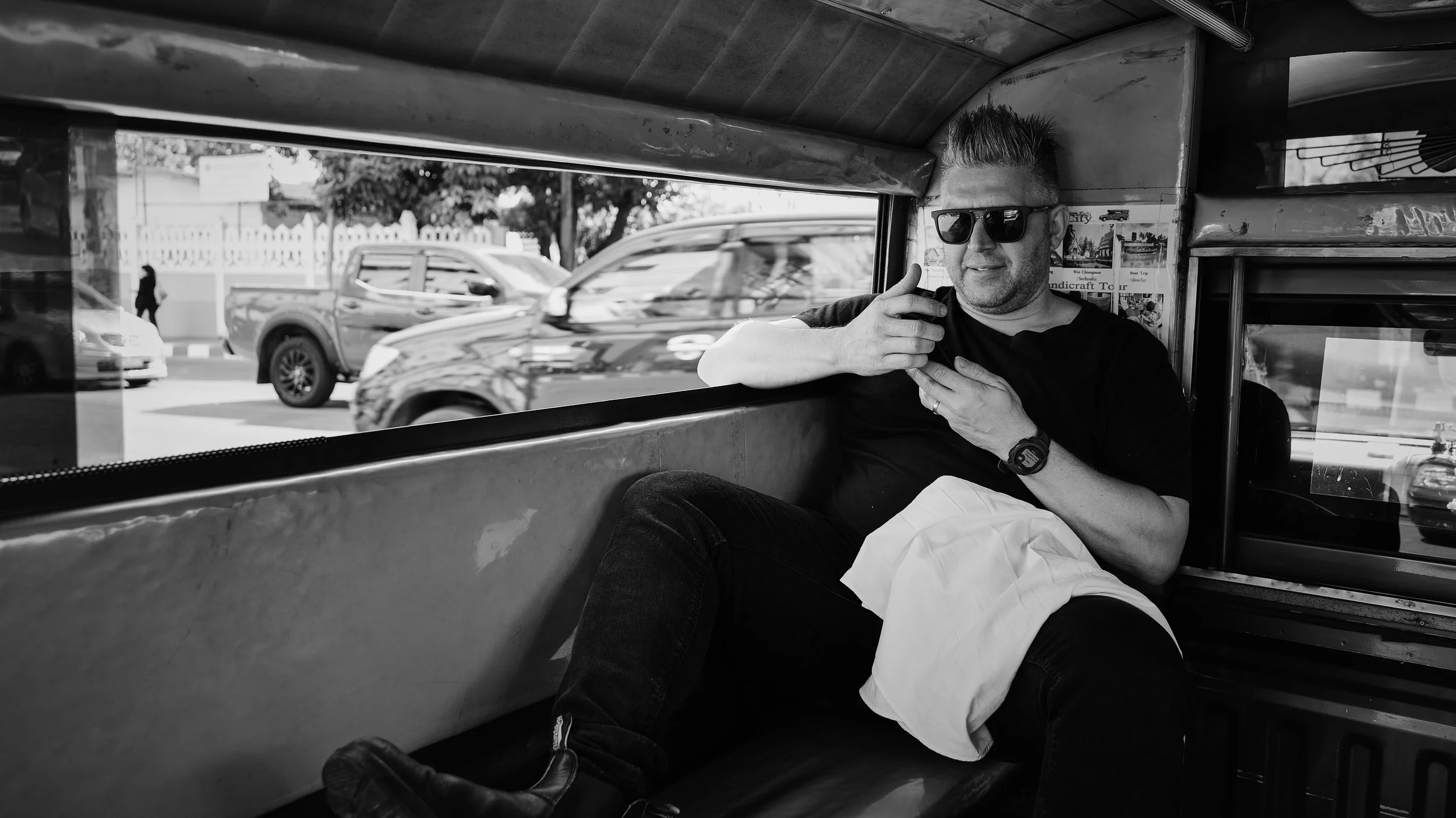 A man with sunglasses, a watch, and a ring, sitting inside a vehicle, smiling, with cars and a pedestrian walking outside visible through the window.