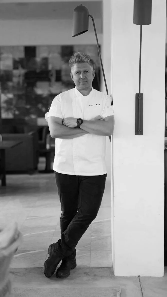 A man wearing a chef's uniform standing with arms crossed in a restaurant, leaning against a white pillar, with a modern interior background.