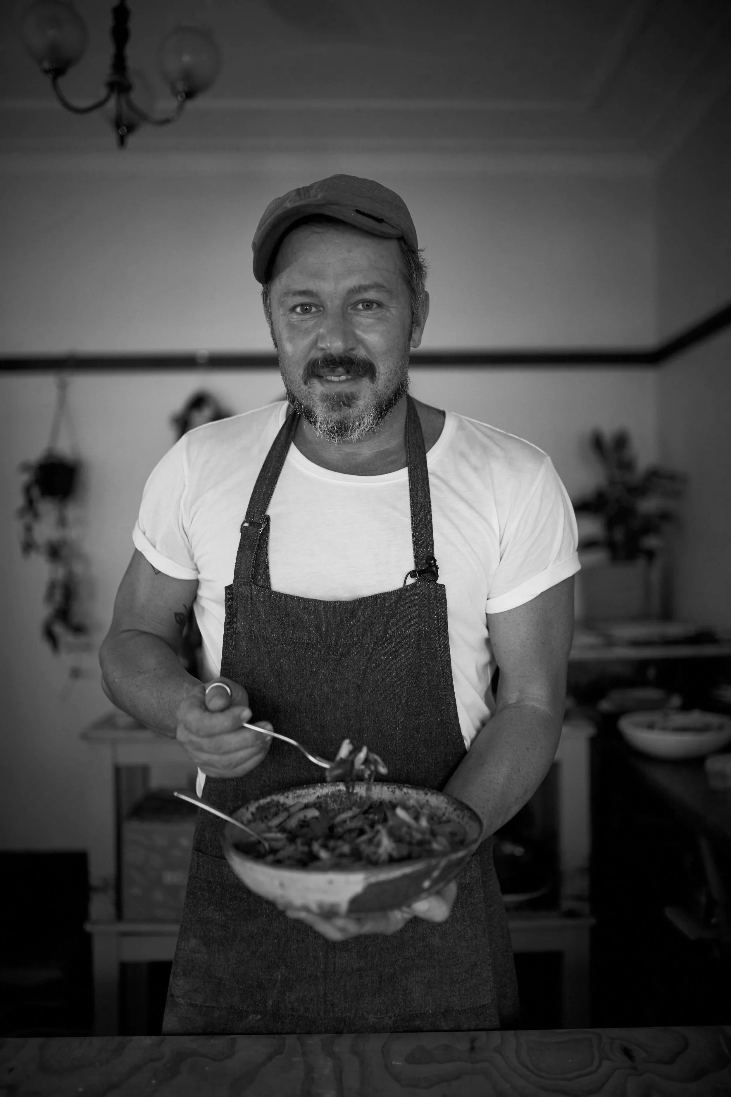 A man with a beard and mustache wearing a cap, t-shirt, and apron, holding a bowl of food and serving it with a spoon in a kitchen.