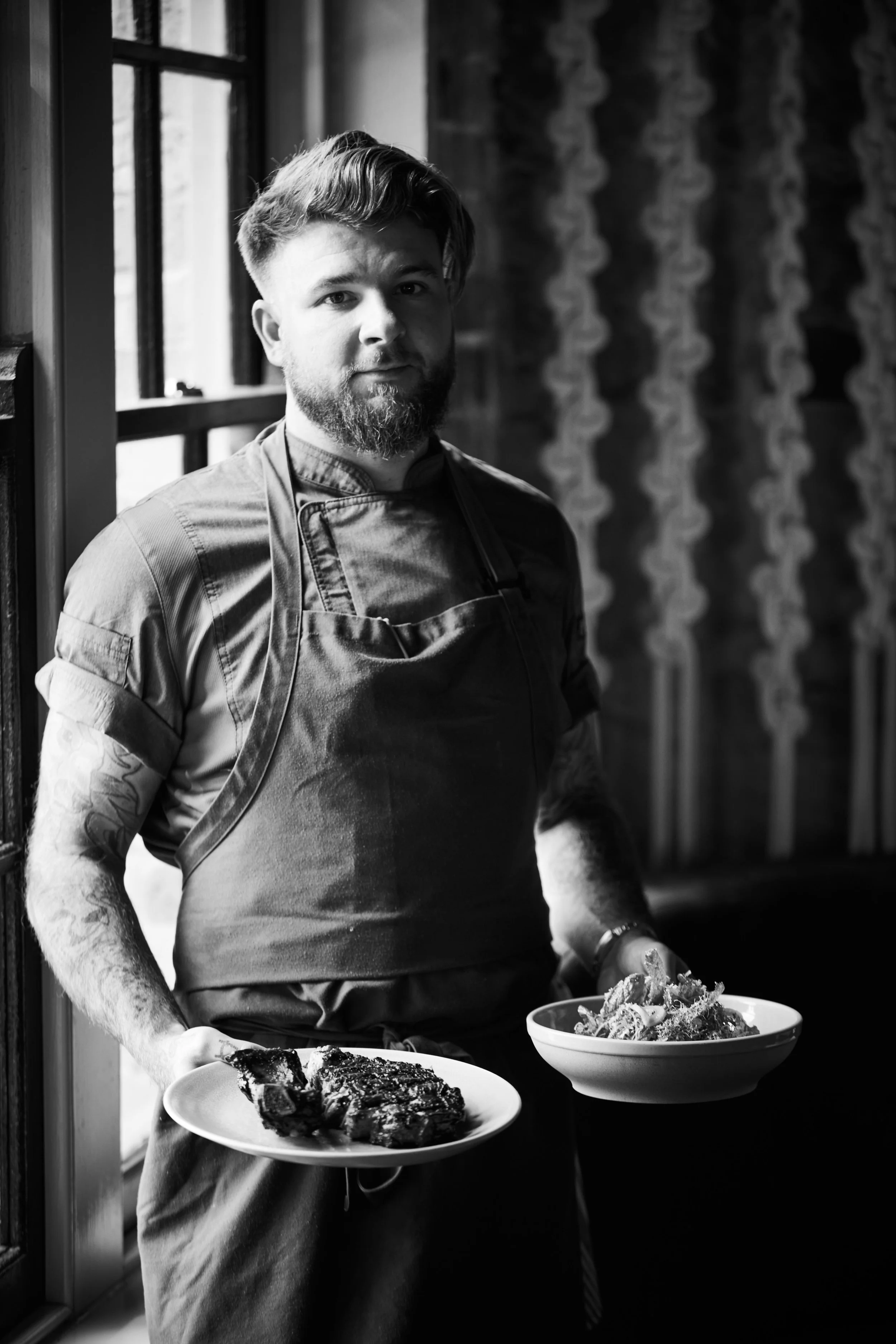A man with a beard and tattoos, wearing a chef's apron, stands by a window in a kitchen, holding plates of food. The interior has a rustic style with patterned wallpaper.