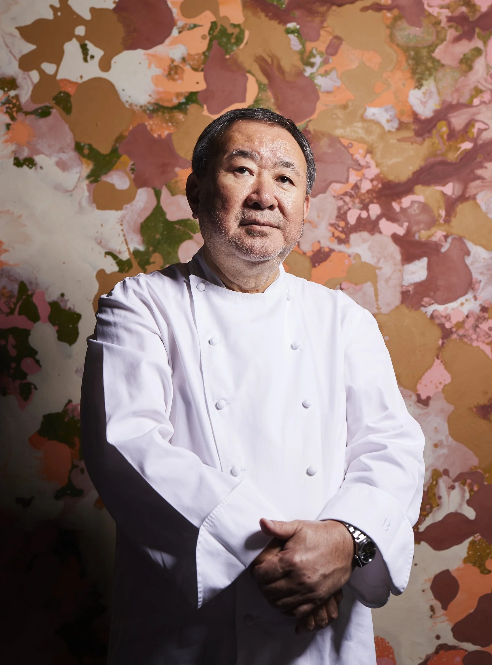 A professional male chef in a white chef's coat standing with arms crossed in front of a colorful abstract background.
