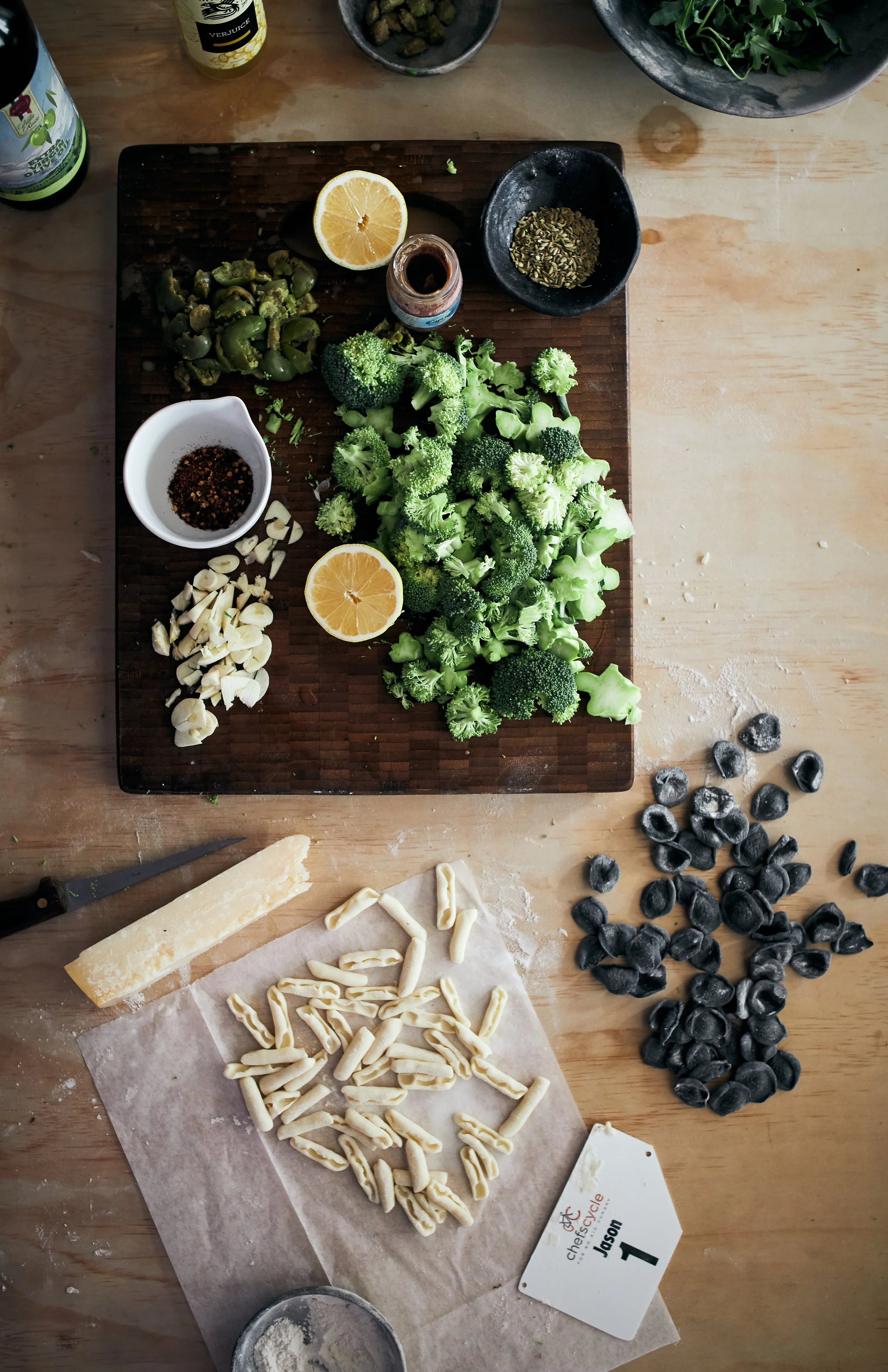 A wooden table with a cutting board holding chopped garlic and a block of Parmesan cheese. To the right of the cutting board are uncooked pasta shapes, and to the upper right, black blueberries. On the table are various ingredients, including broccoli florets, lemon halves, chopped green vegetables, dried herbs, crushed red pepper, and bottles of beverages.