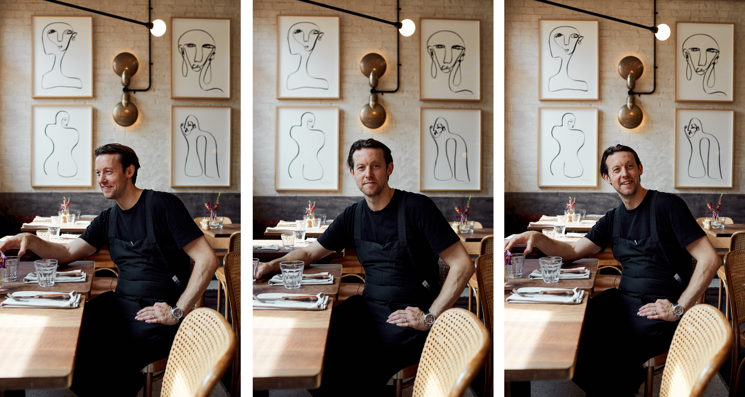 A man wearing a black shirt and apron, seated at a restaurant table with glasses and napkins, smiling and relaxing. Behind him is a brick wall decorated with abstract line art portraits of faces framed and two round wall lamps.