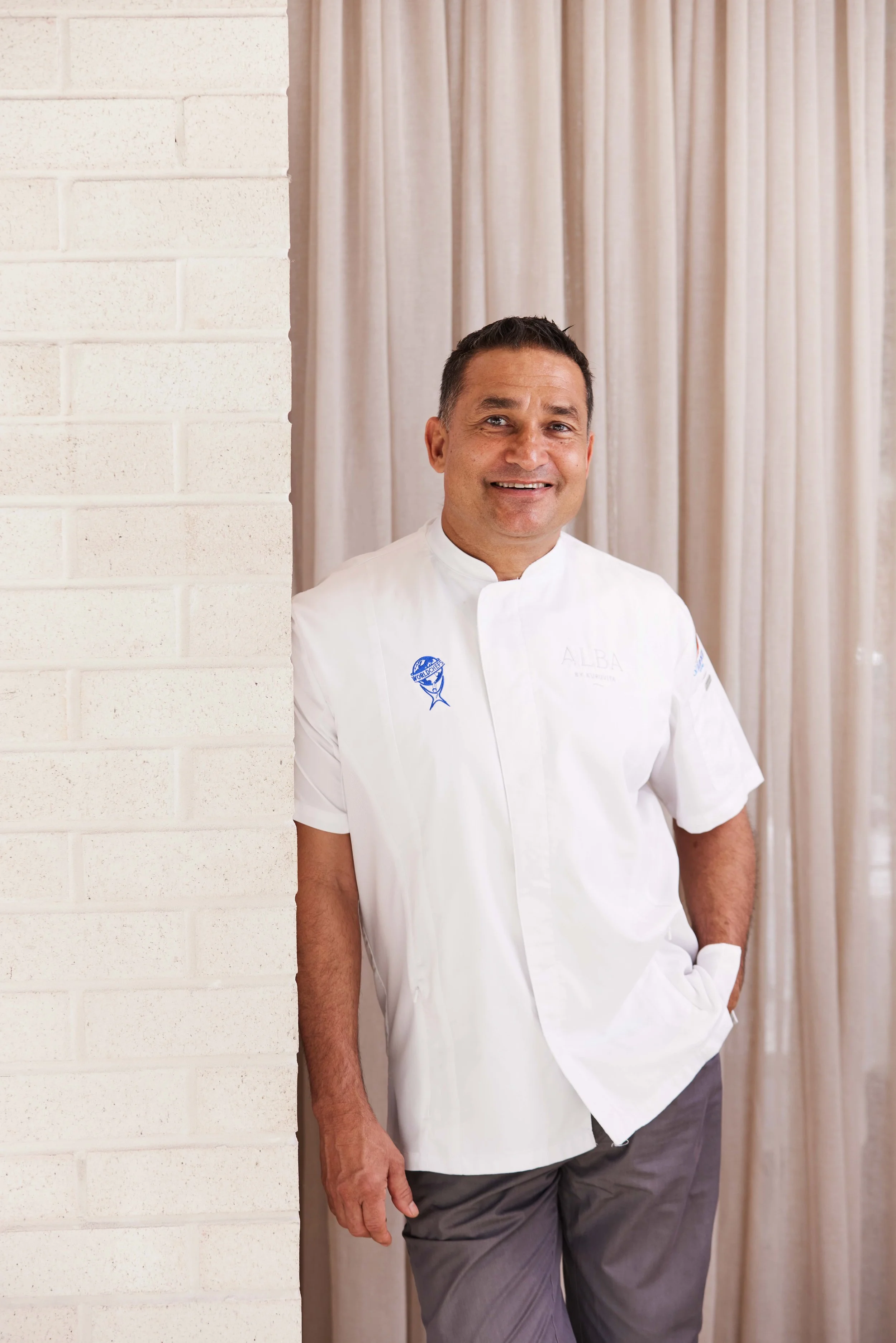 A man in a white chef's coat and gray pants stands in front of cream-colored curtains, smiling at the camera.