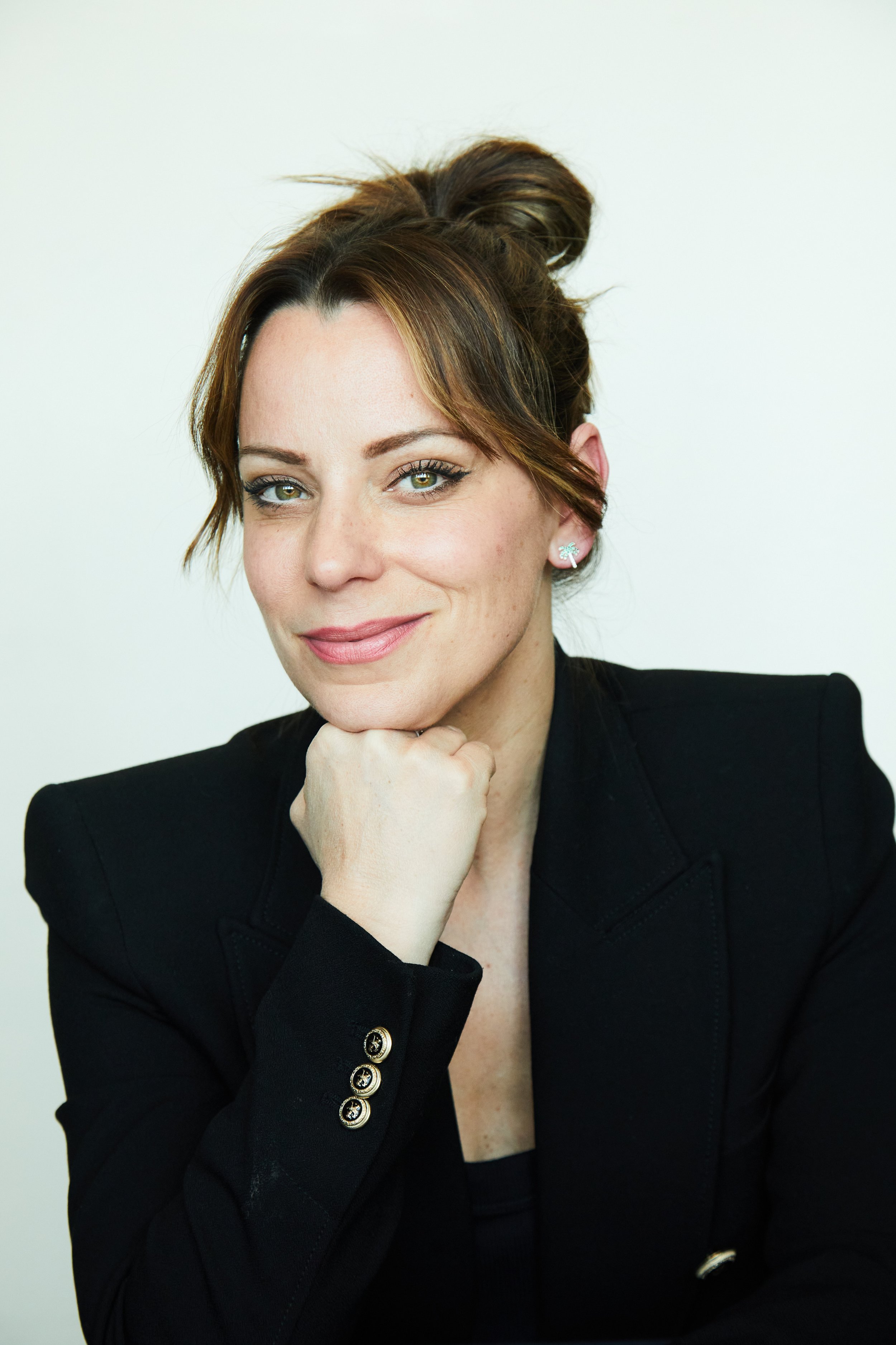 A woman with brown hair styled in a top bun, wearing a black blazer with gold button details, and earrings shaped like a starfish, resting her chin on her hand and smiling softly against a plain white background.